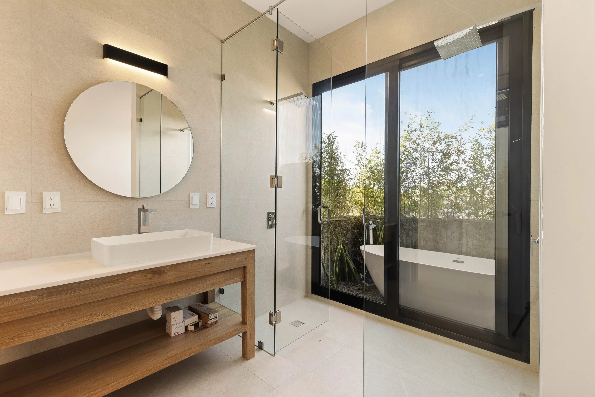 Modern bathroom with a round mirror, a vessel sink on a wooden vanity, a glass shower enclosure, and floor-to-ceiling windows overlooking greenery with a freestanding bathtub outside.