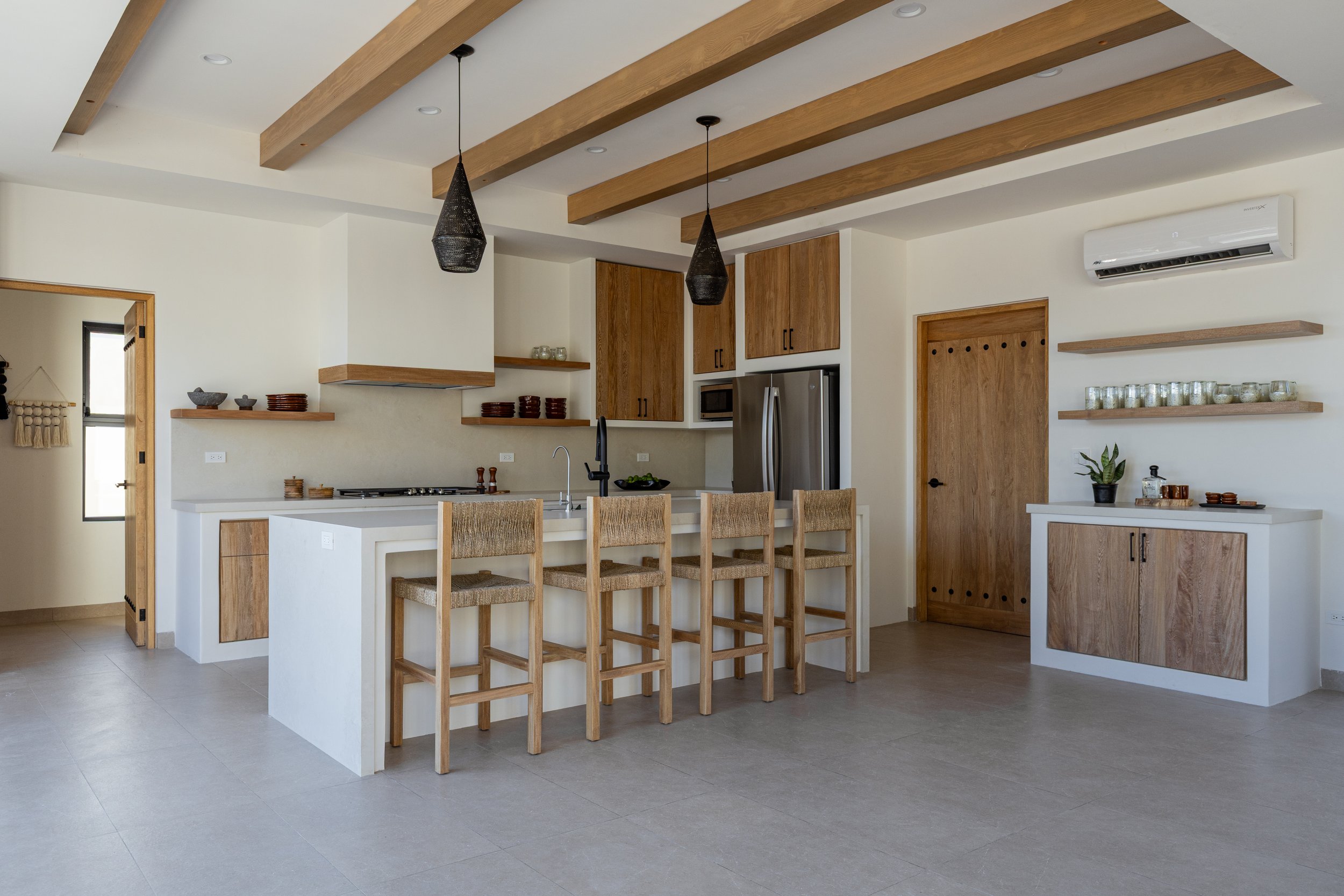 Modern kitchen with white and wood cabinetry, a kitchen island with four wooden bar stools, black pendant lights, a stainless steel refrigerator, microwave, and a white air conditioning unit on the wall.