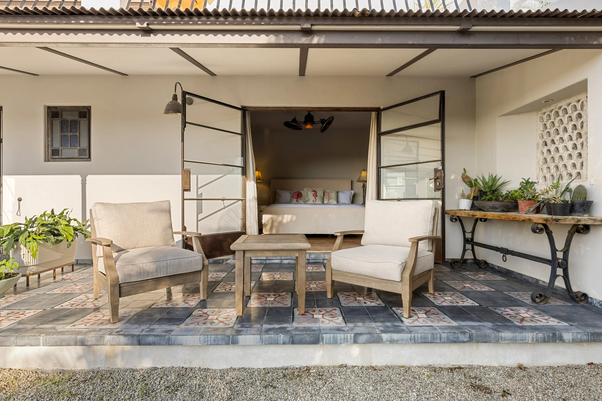 A cozy outdoor patio with two beige armchairs and a small wooden table, leading into a bedroom with a large bed and bedside lamps, decorated with greenery and potted plants on shelves, set on patterned tile flooring.
