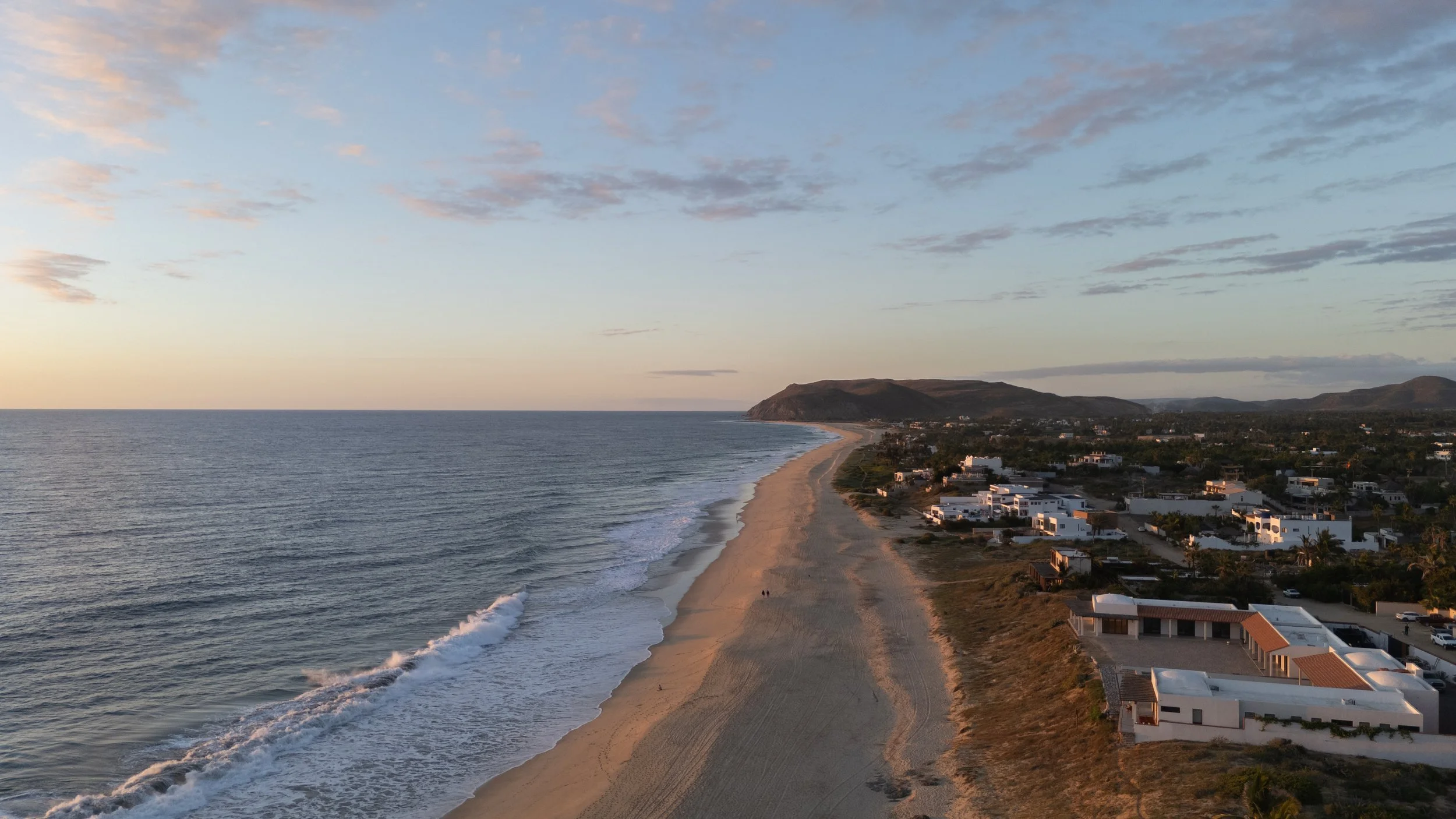 Aerial view of a beach with houses along the coast, ocean waves, and hills in the background during sunset.