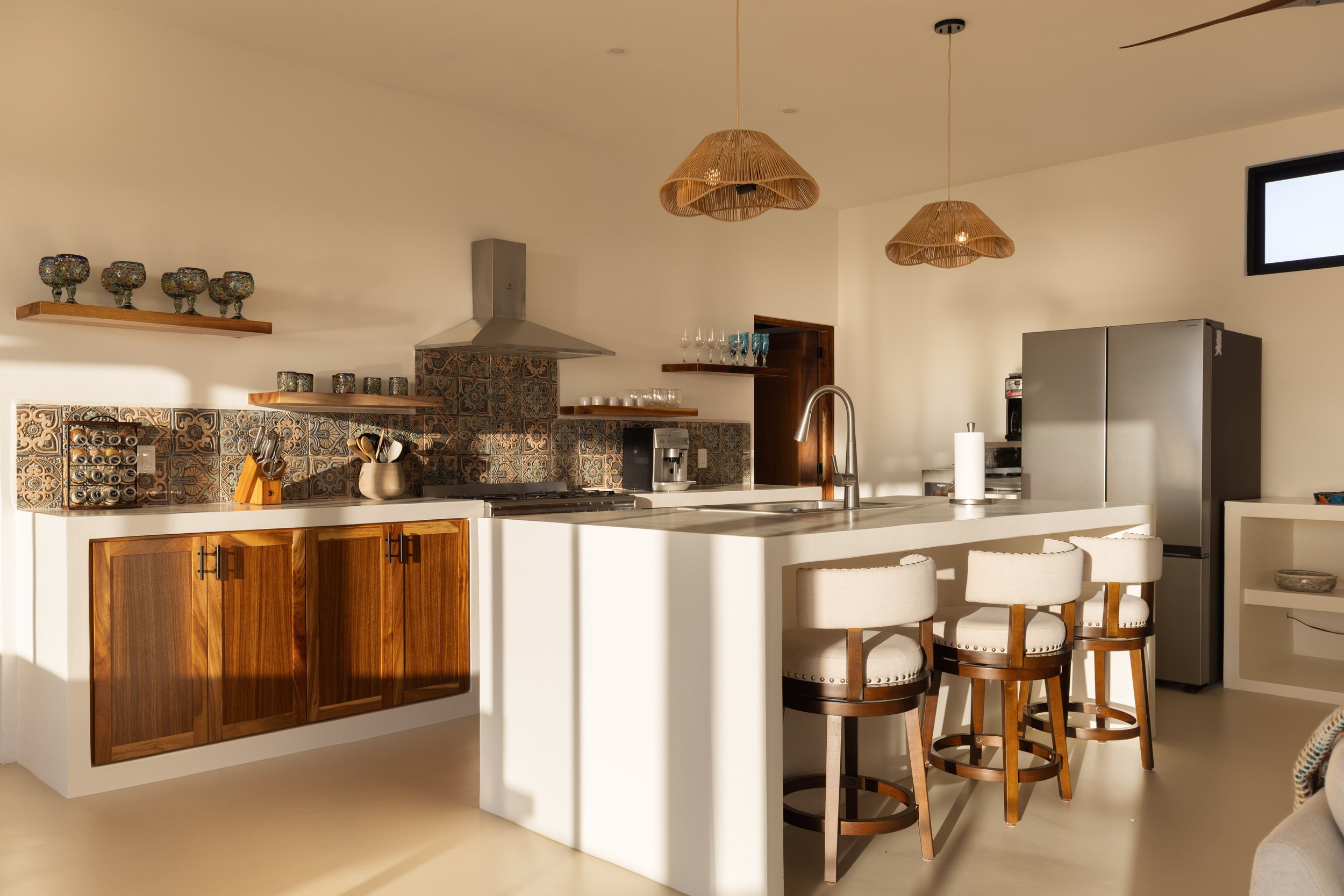 Modern kitchen with white countertops, wooden cabinets, patterned tile backsplash, three white barstools, stainless steel refrigerator, and pendant lights.