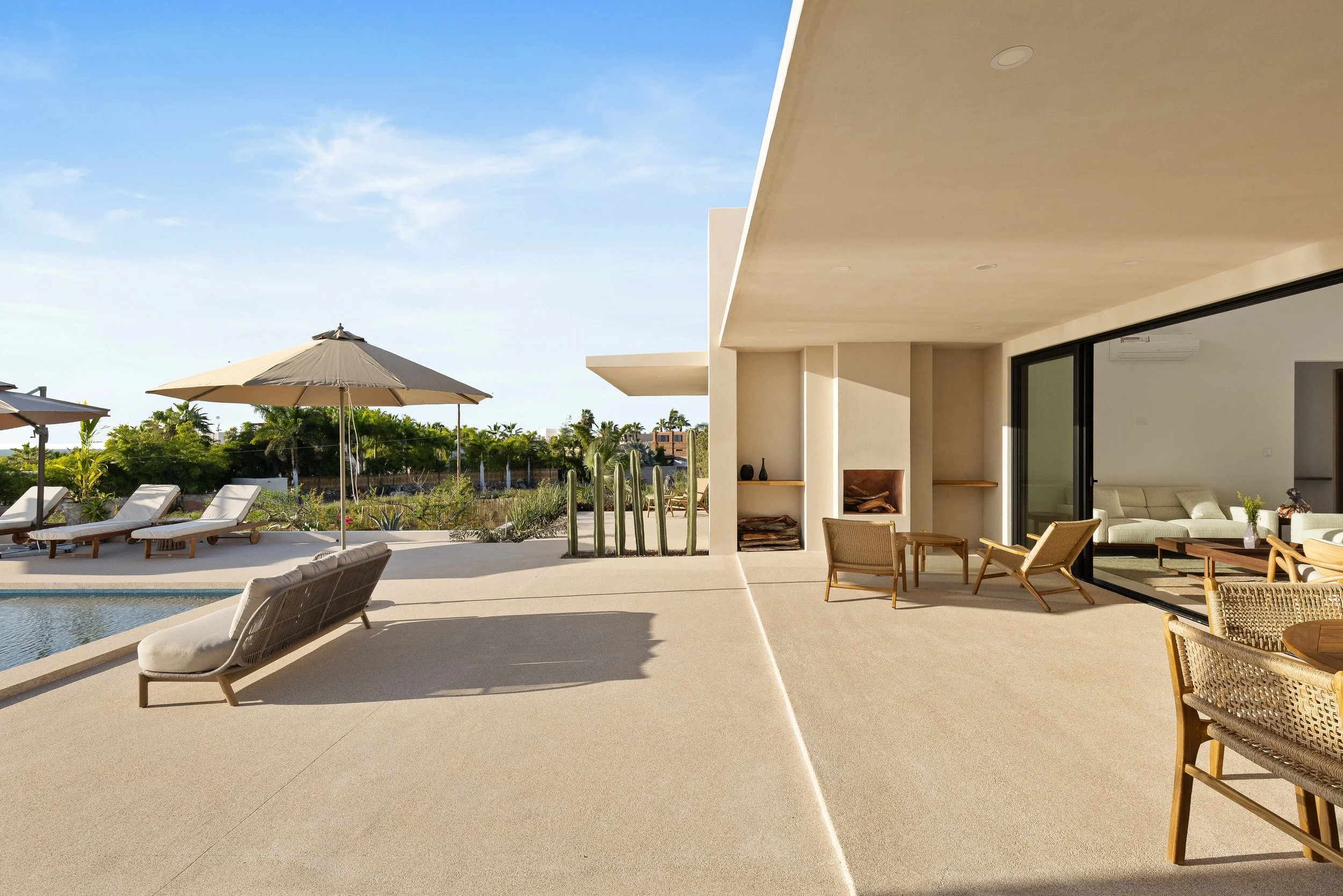 Modern outdoor patio with lounge chairs, umbrellas, and view of greenery and blue sky, connected to a sleek indoor living room with sliding glass doors.
