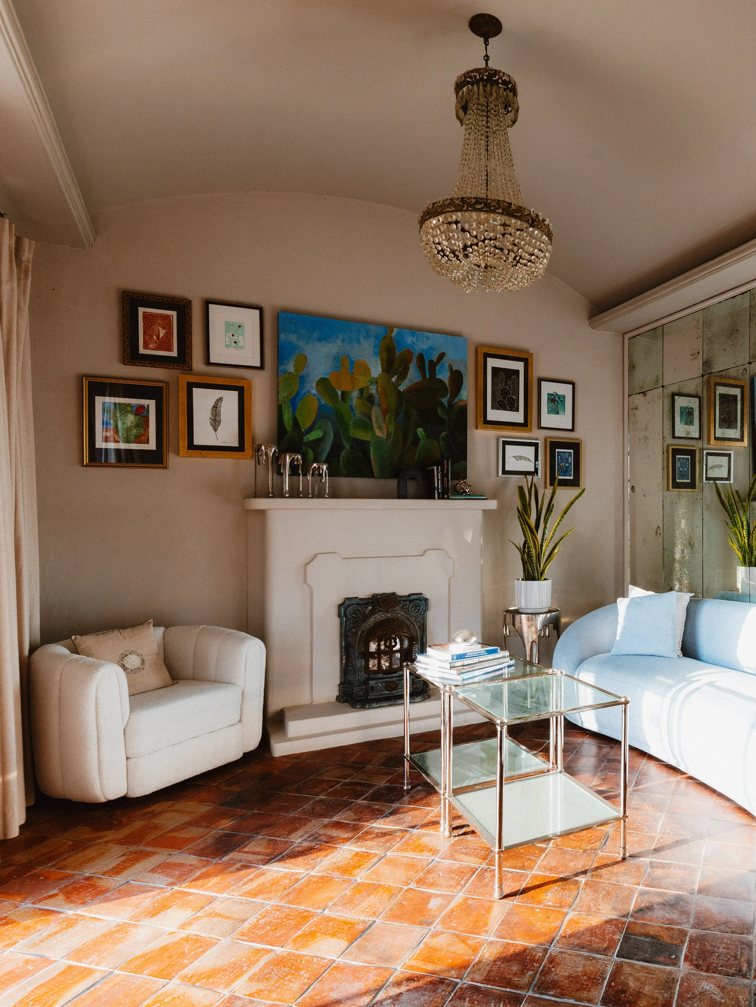 Living room with white sofa, white armchair, glass coffee table, artwork on wall above white fireplace, chandelier, potted plants, and tiled floor.