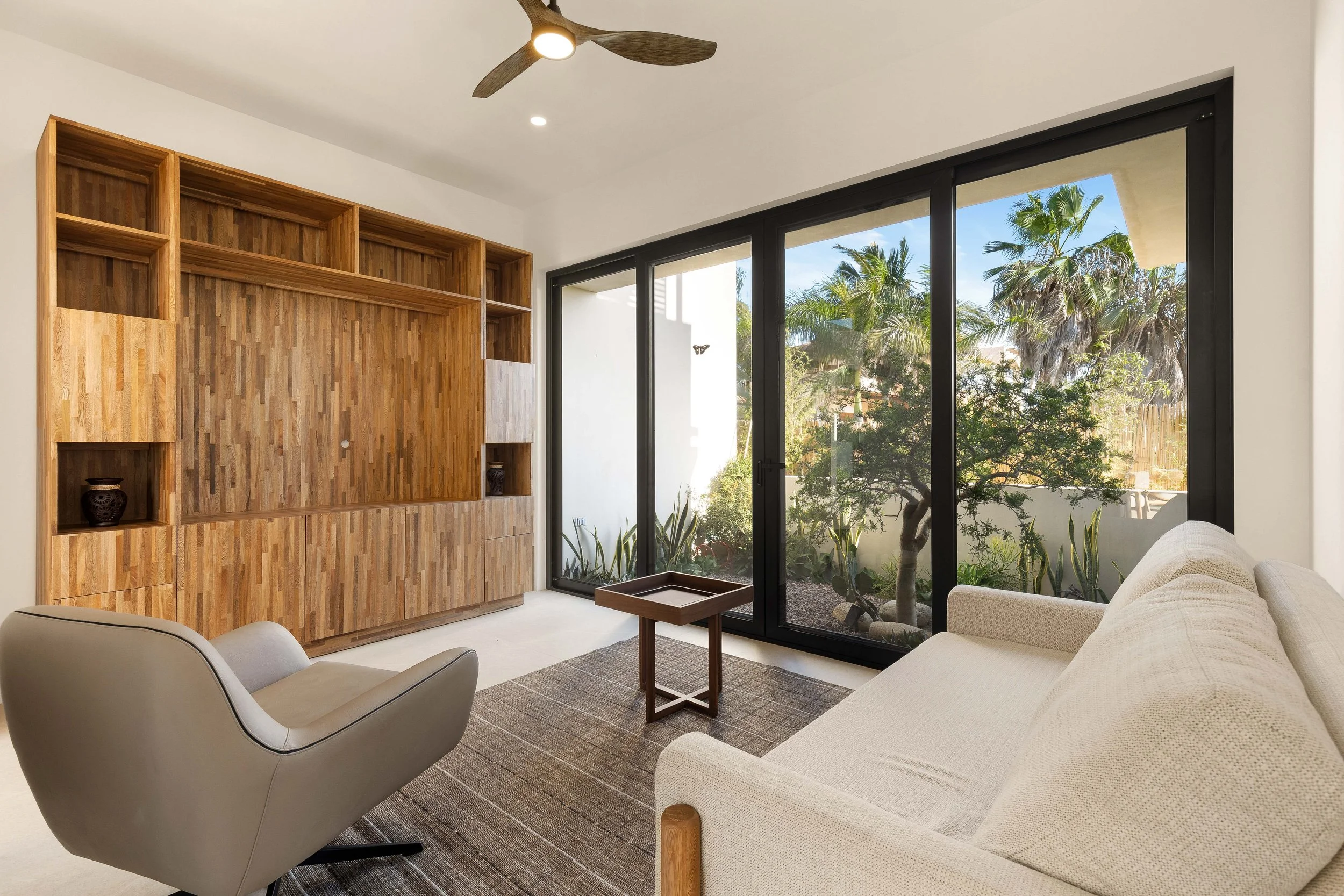 Living room with a beige sofa, gray armchair, wooden shelving unit, glass sliding doors, and garden view with trees.