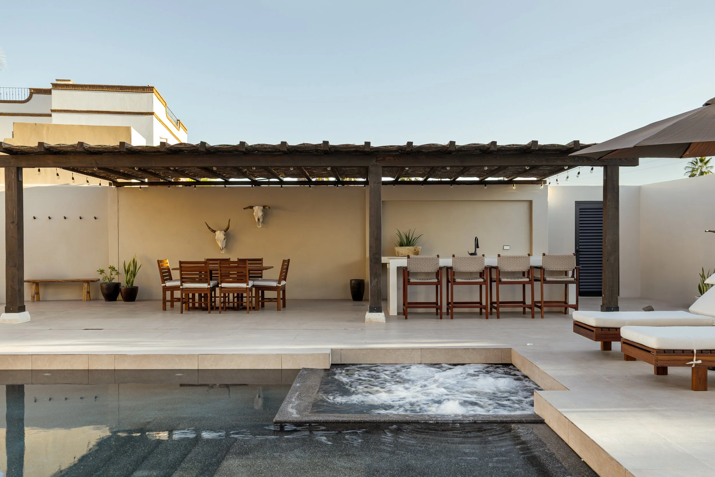Outdoor patio with a pool in the foreground, a shaded dining area with wooden table and chairs, bar stools at a counter, and decorative skull and bull head wall art, surrounded by white walls and potted plants.