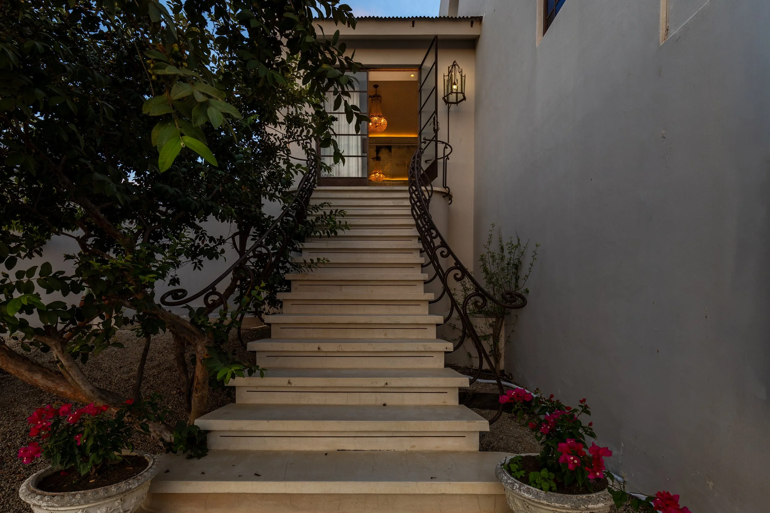 Exterior staircase with marble steps, ornate iron railings on both sides, leading to a door with glass panels inside a building. Potted flowers with pink blooms sit at the base of the stairs, and a tree with green leaves is on the left side.