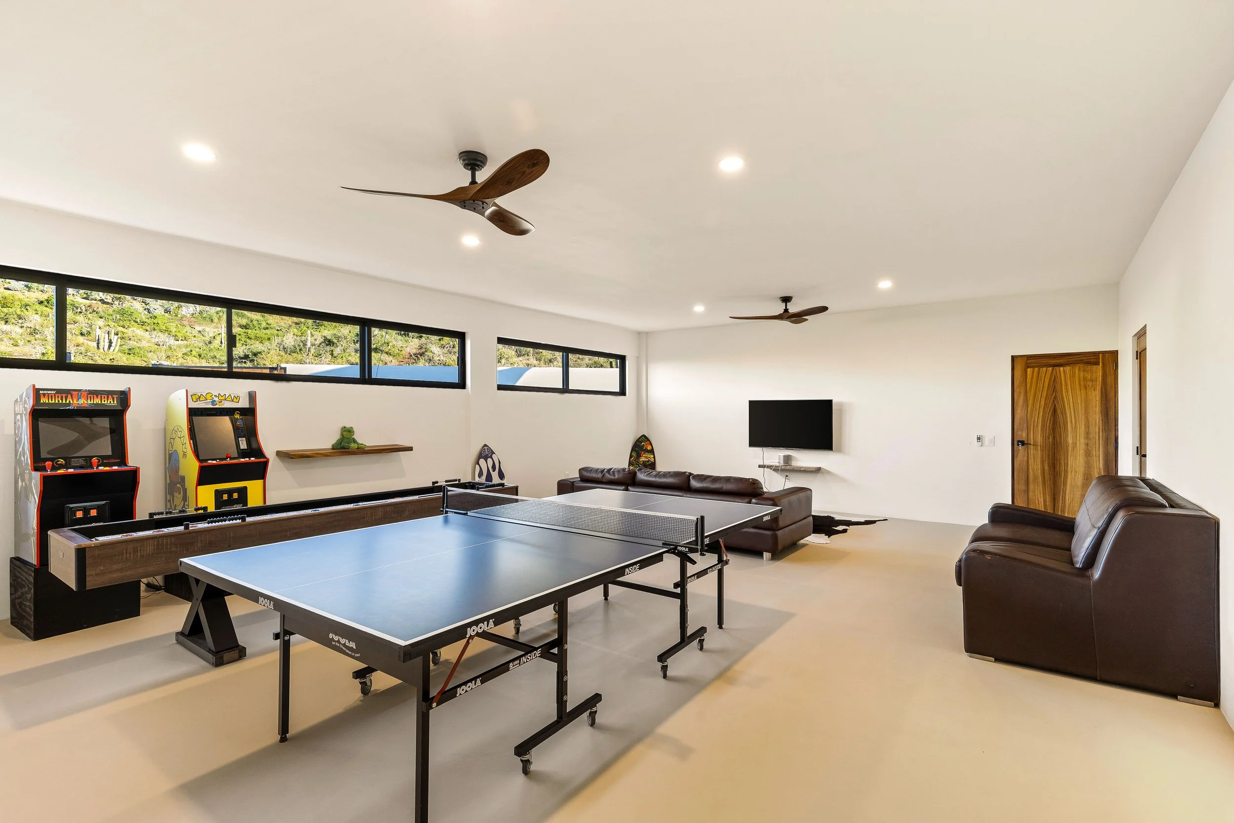 Spacious game room with ping pong table, two arcade machines, wall-mounted TV, leather sofas, ceiling fans, and windows showing greenery outside.