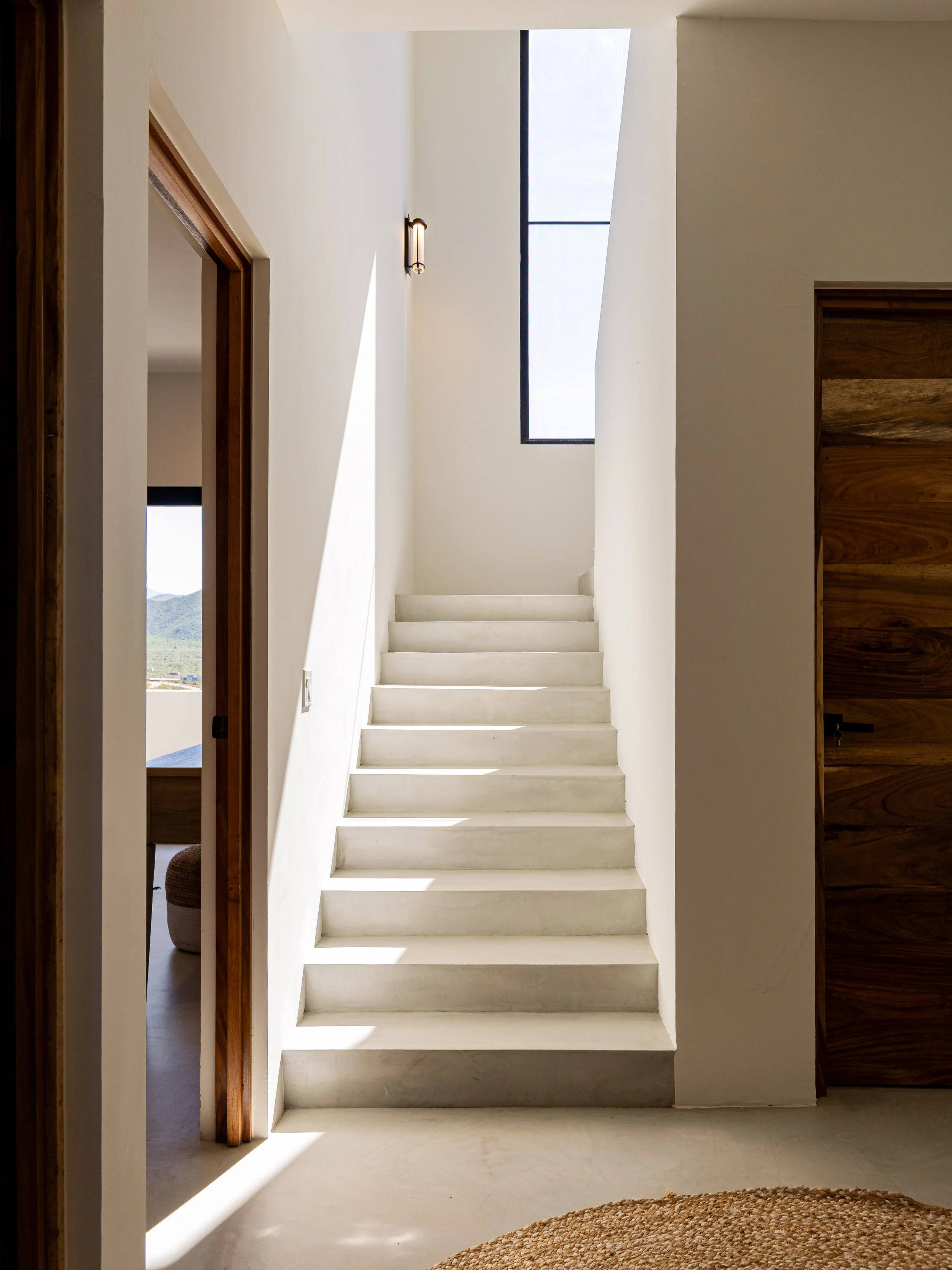 Interior view of a modern home with a staircase leading to the upper floor, illuminated by natural light coming through a tall window.