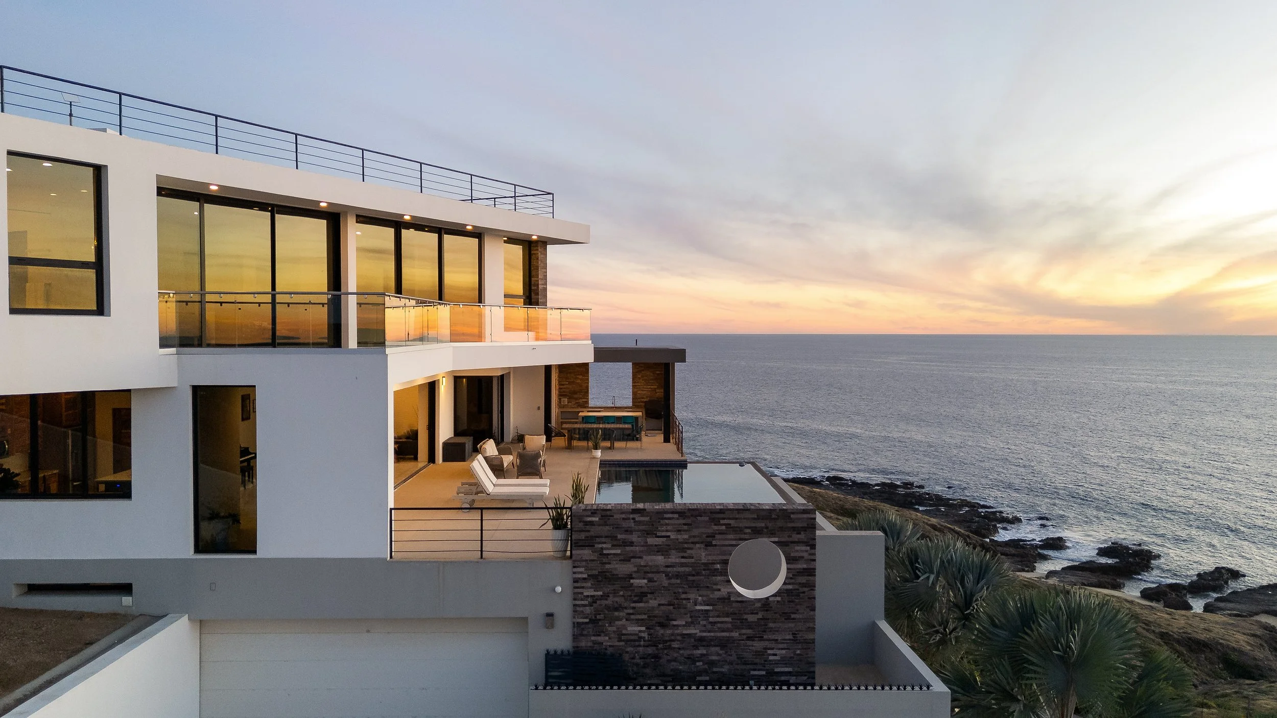 Modern beachfront house with large glass windows and outdoor patio, overlooking the ocean at sunset.