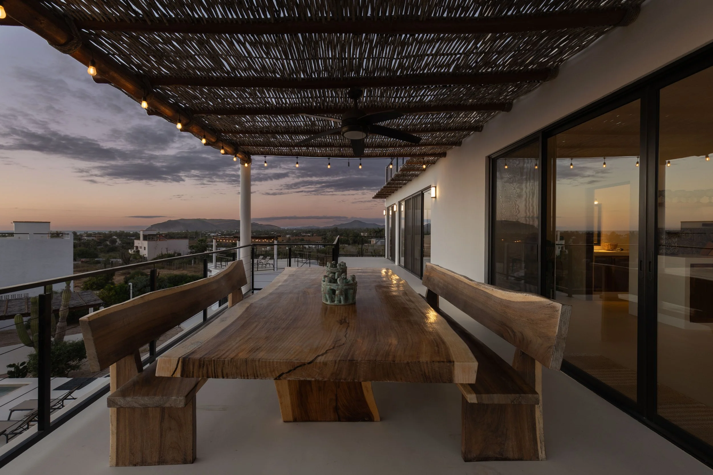 Outdoor balcony with a wooden dining table and benches, string lights overhead, and a view of a sunset over a desert landscape with mountains in the distance.
