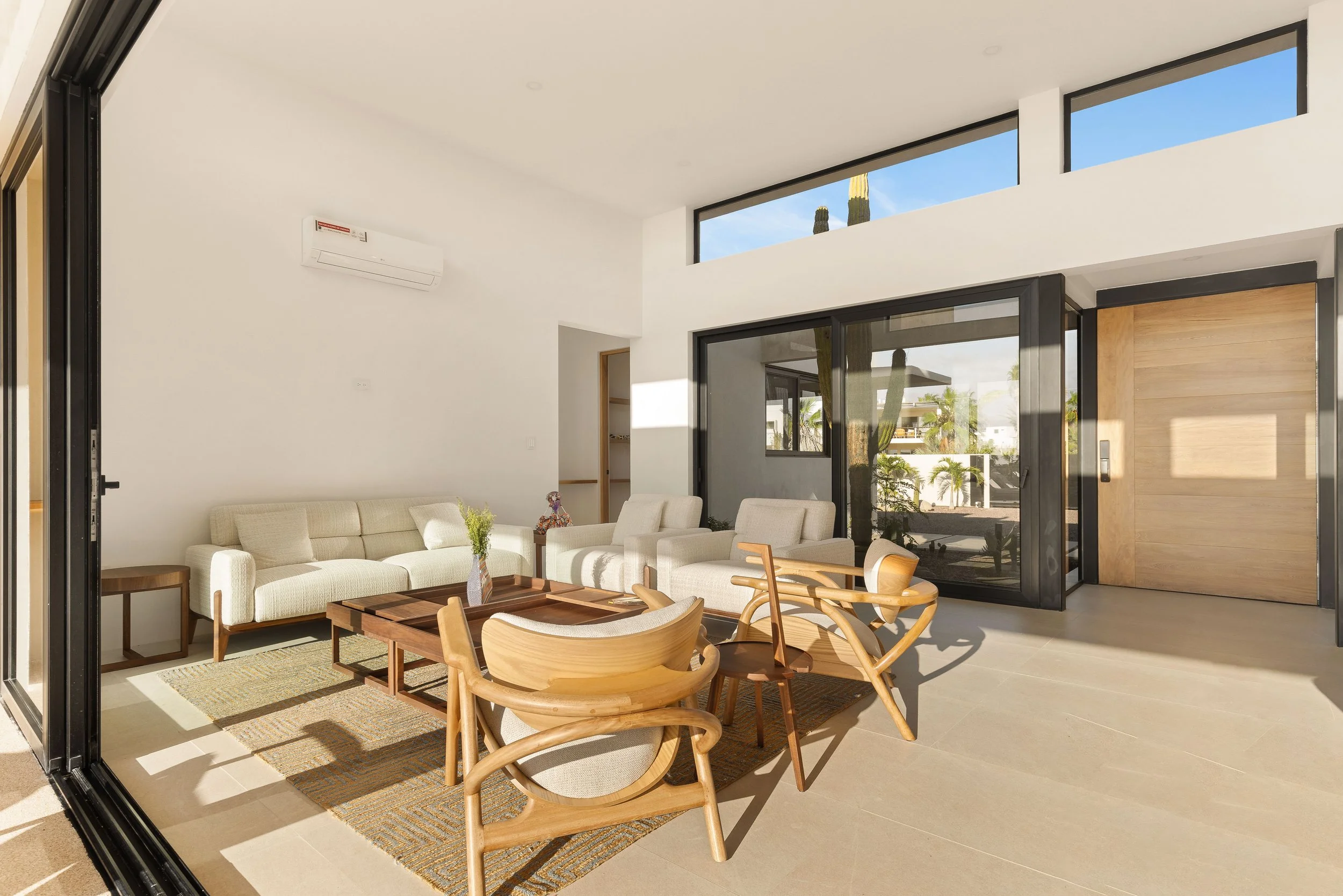 Modern living room with white walls, beige sofas, wooden chairs, a coffee table, and large glass sliding doors leading to outdoor space with cacti and palm trees.