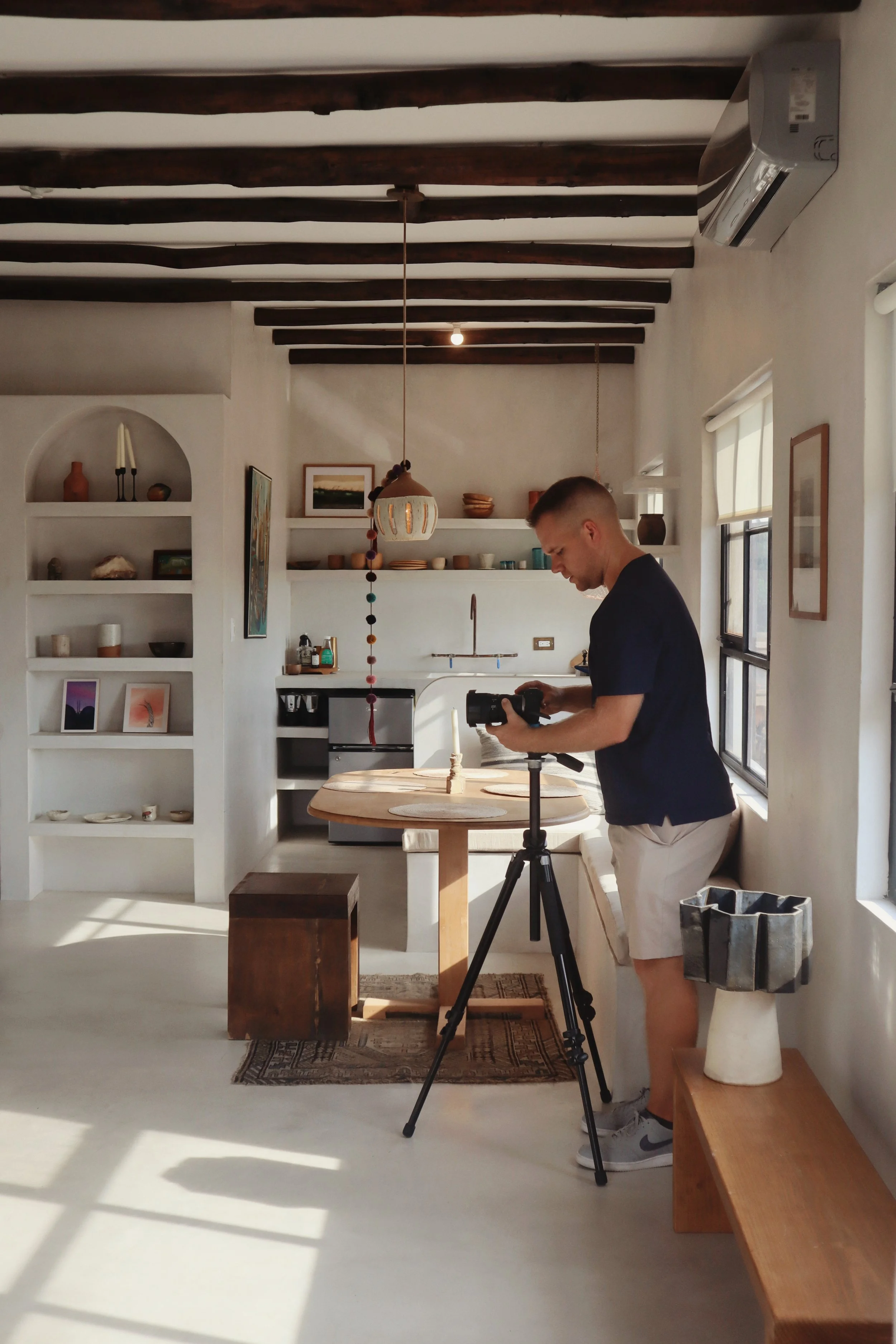 A man filming in a bright, modern kitchen with wooden beams on the ceiling, white walls, and shelves with decorative items. He stands on a rug near a round dining table, adjusting a camera on a tripod.