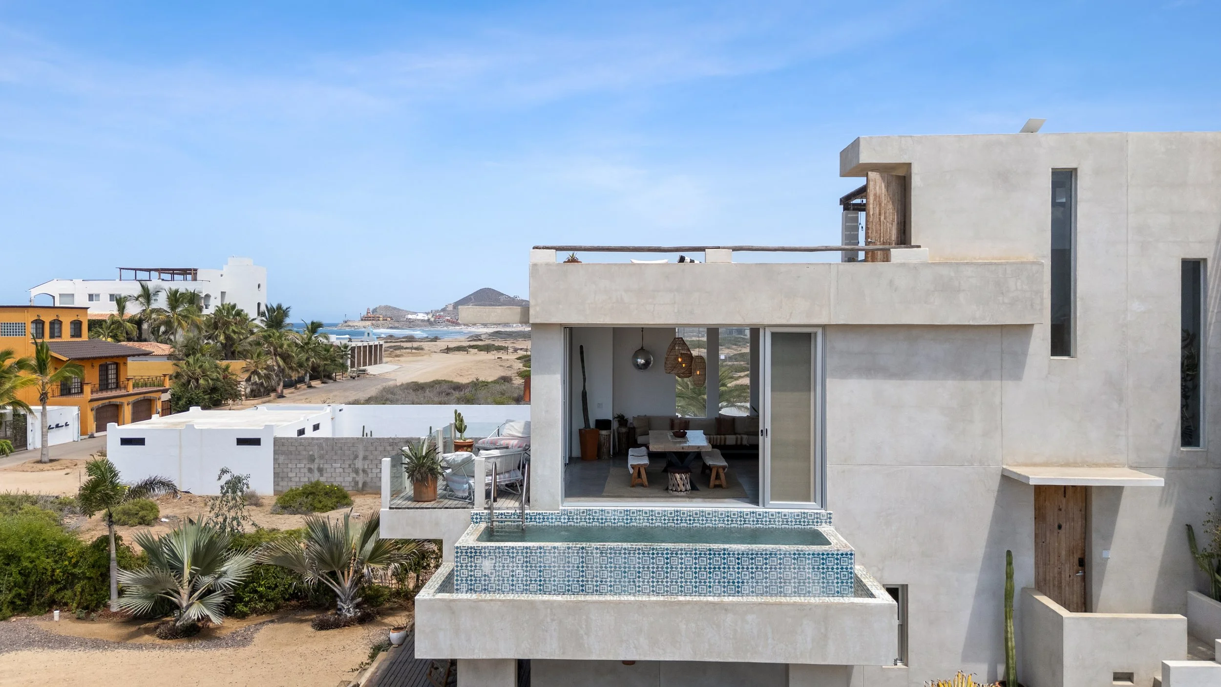 Modern white house with outdoor balcony featuring an infinity pool, desert landscape, palm trees, neighboring colorful houses, and mountains in the background.