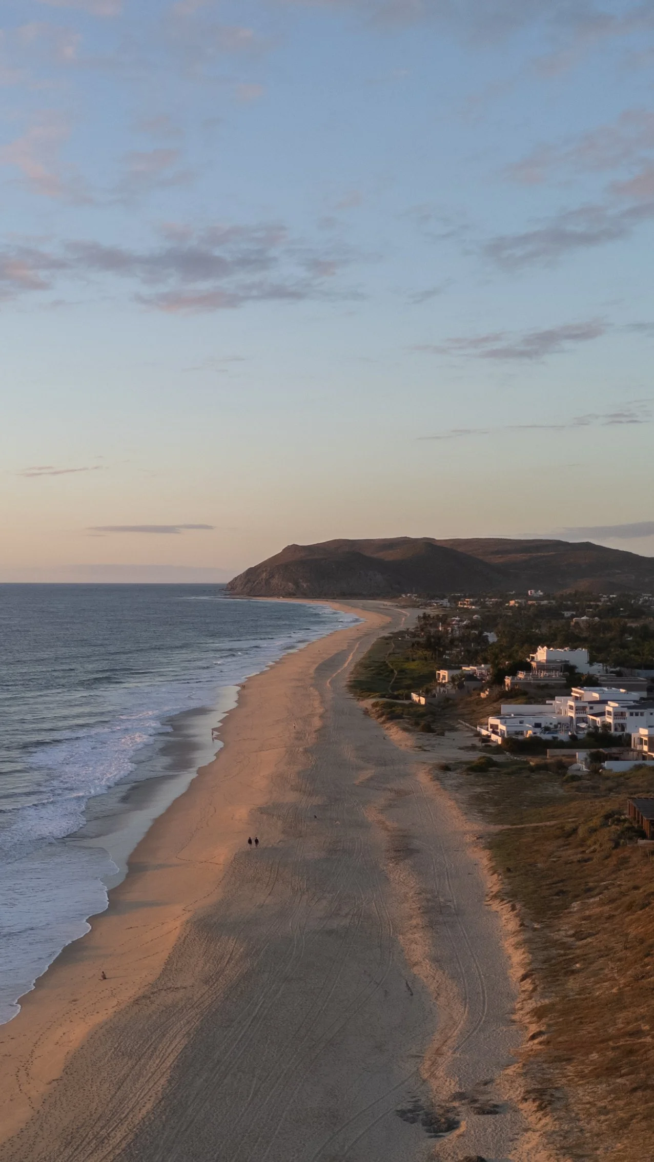 Aerial view of a coastline with a sandy beach, ocean waves, residential buildings, and a hilly landscape in the background during sunset.