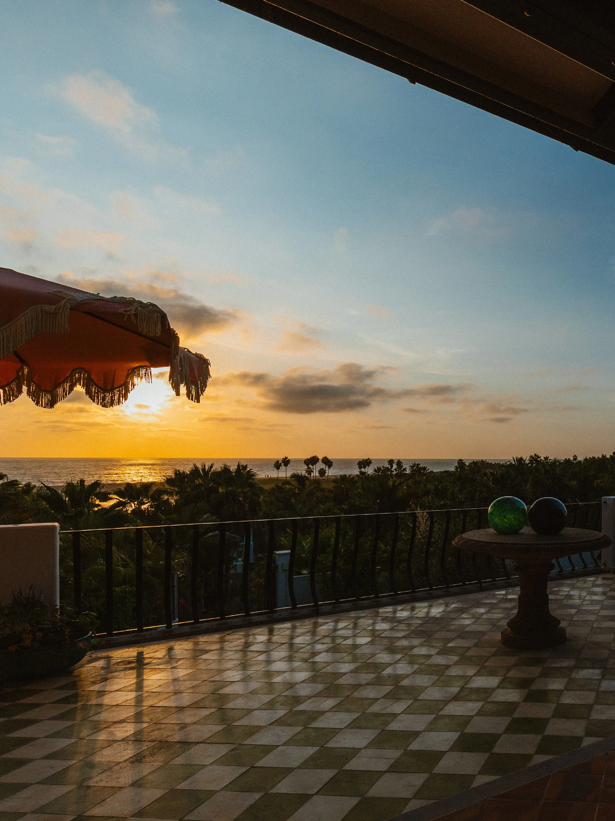 A balcony overlooking trees and the ocean at sunset, with a red and fringed umbrella on the left and two glass balls, one green and one black, on a small table on the right.