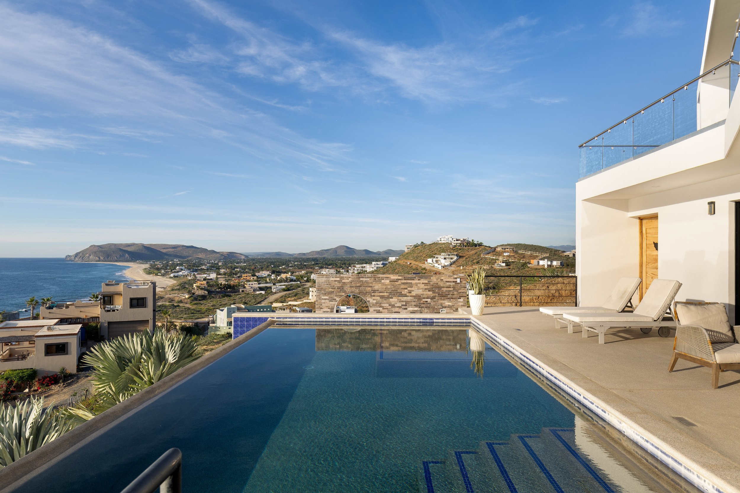 A scenic view of a rooftop infinity pool with lounge chairs, overlooking a coastal landscape with hills, beach, and ocean under a blue sky.