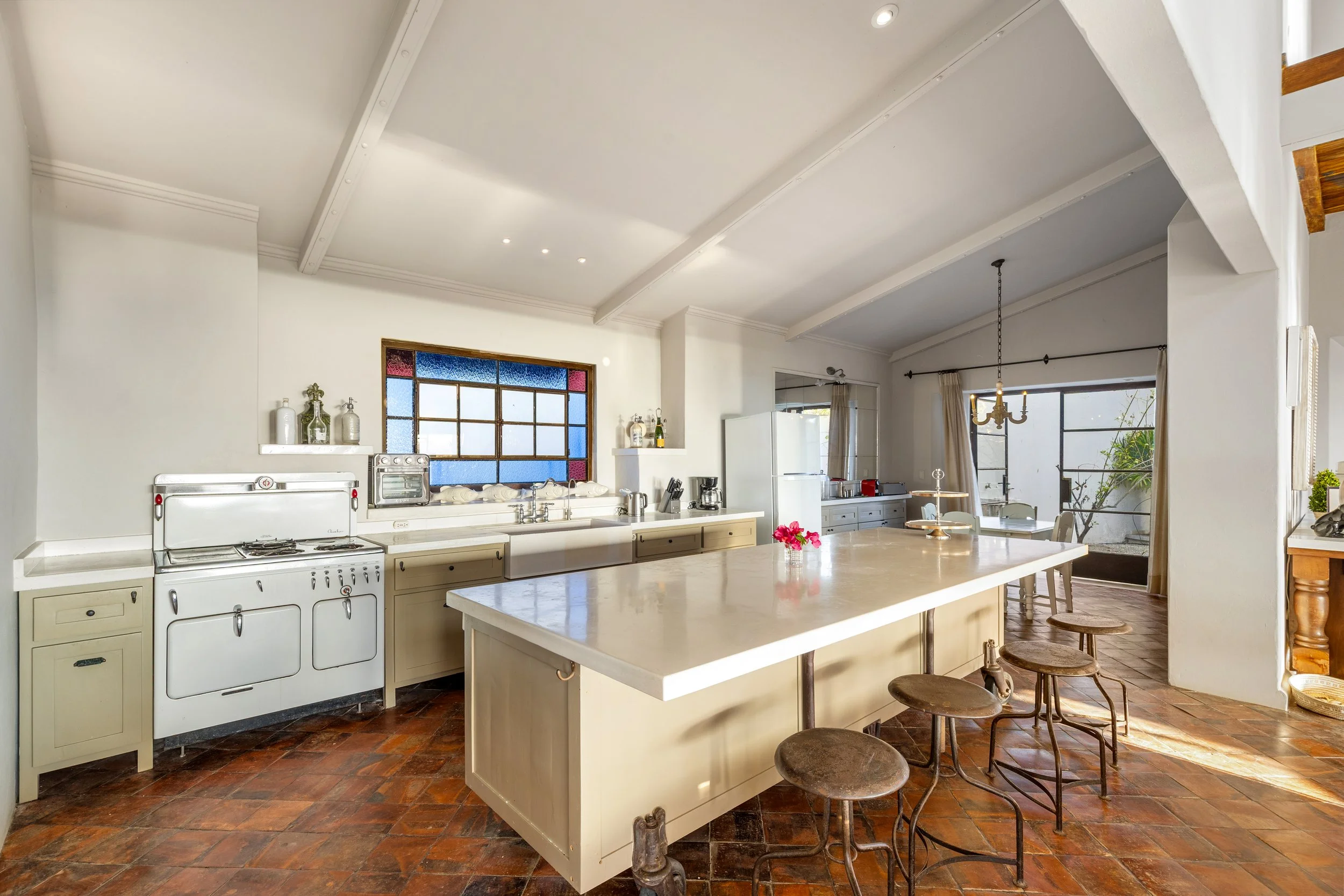Bright kitchen with white cabinets, vintage stove, large white island, wooden bar stools, terracotta tile floor, and large windows with curtains.