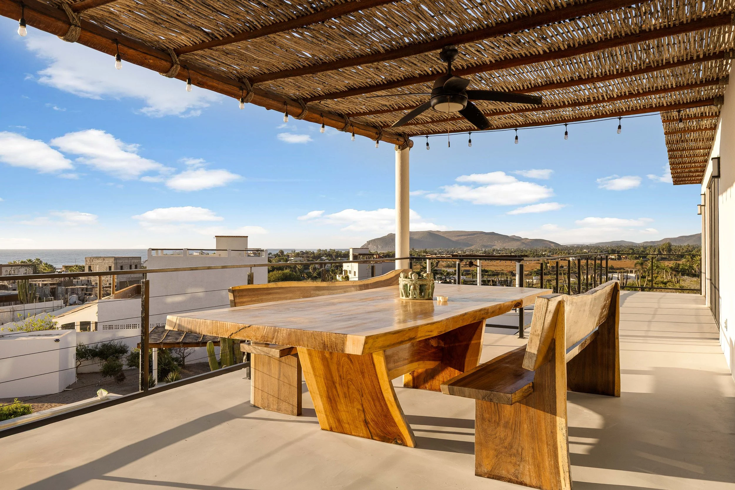 Outdoor patio with a wooden table and benches, overlooking a cityscape and mountains under a blue sky with scattered clouds.