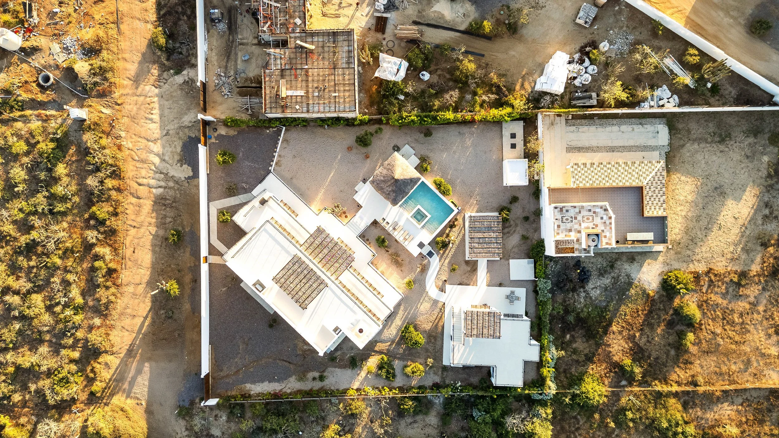 An aerial view of a property with white buildings, a small swimming pool, and a courtyard, surrounded by trees and dry land.