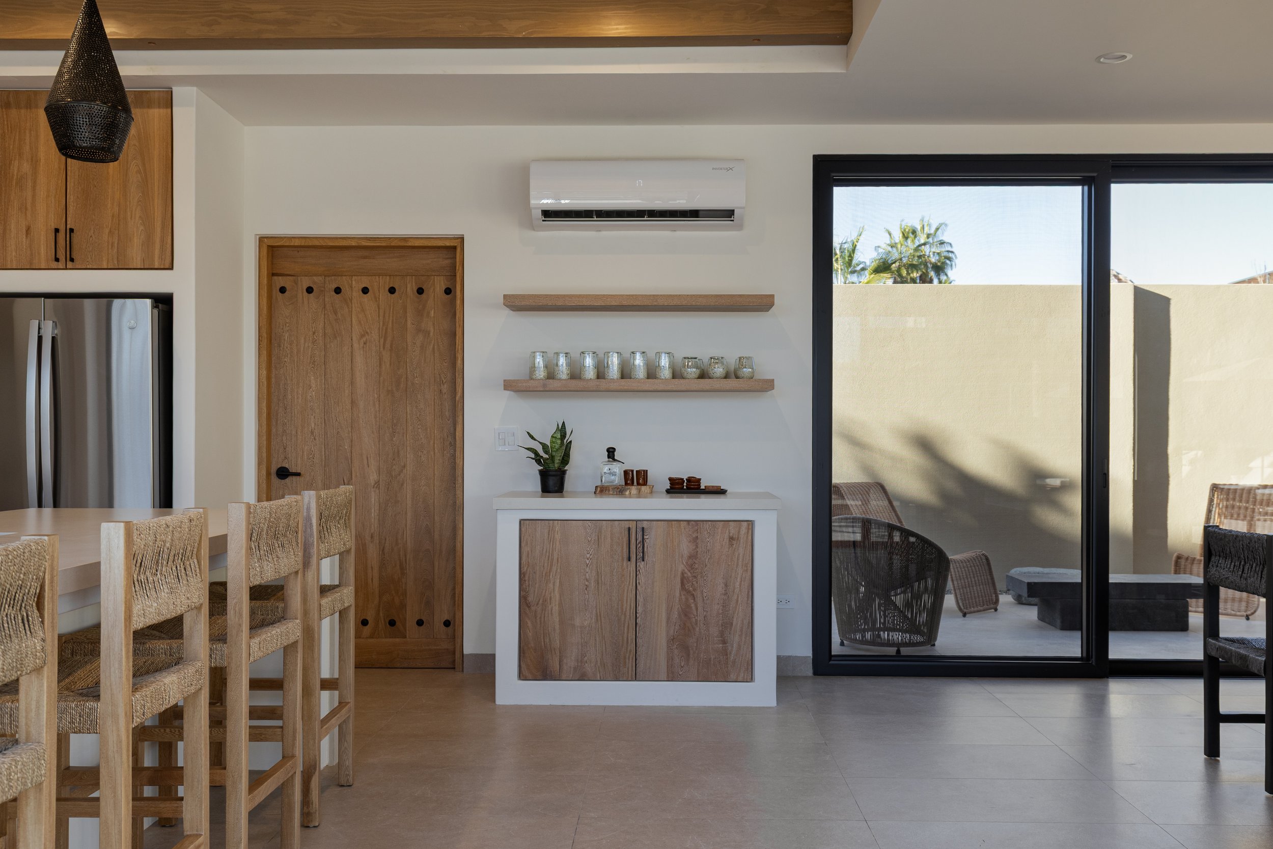Modern kitchen with wooden cabinets, bar stools, a sliding glass door leading to an outdoor patio, a wall-mounted air conditioning unit, and a small white and wood cabinet with decorative items on top.
