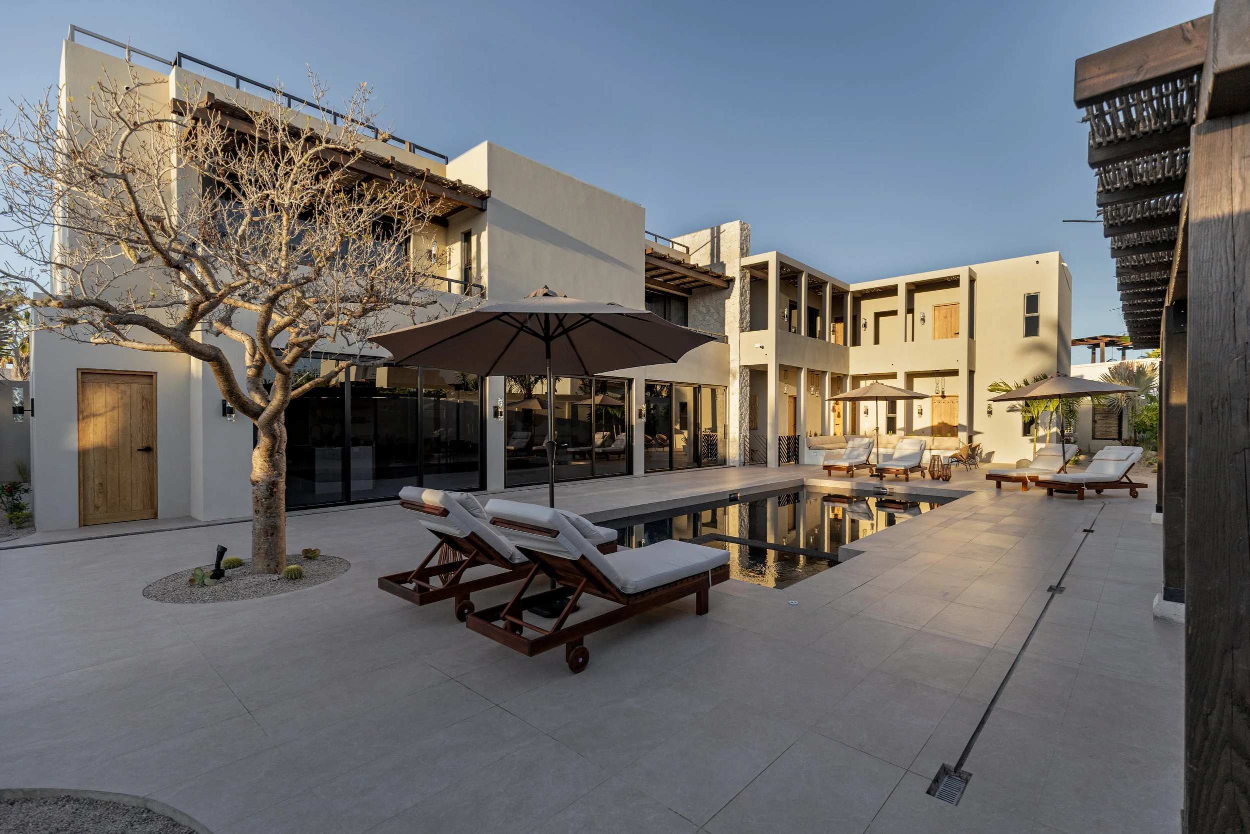 Modern residential backyard with a swimming pool, lounge chairs with cushions and umbrellas, a leafless tree, and a two-story house with balconies and large windows during daytime.