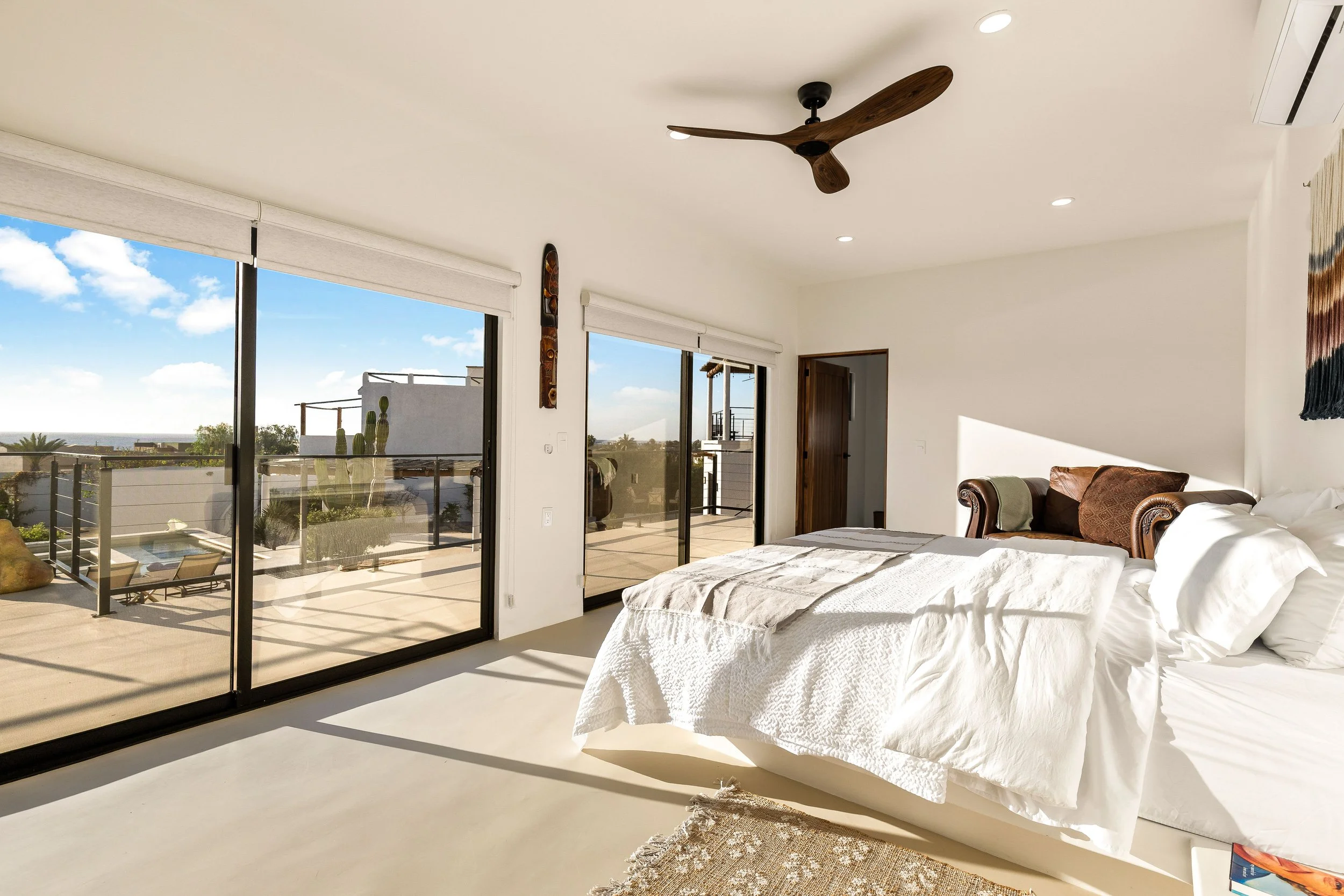A bedroom with large glass sliding doors leading to a balcony, a white bed with pillows, a brown armchair, and a ceiling fan.