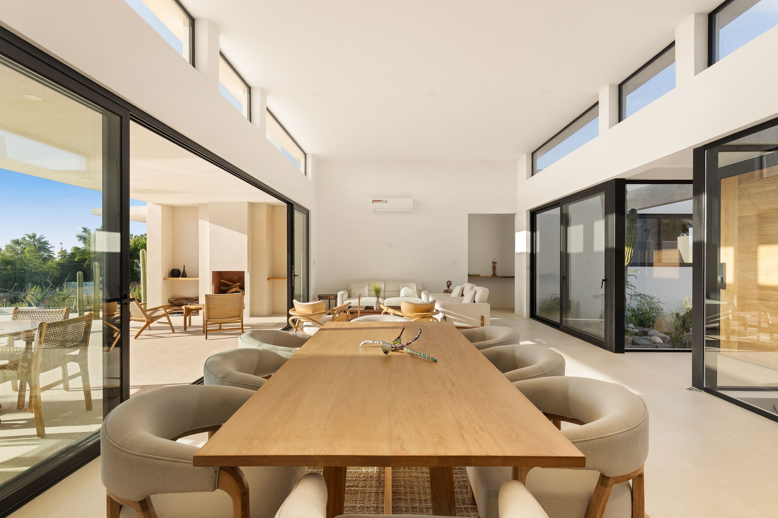 Spacious modern dining and living area with large glass sliding doors, white walls, and minimalist furniture including a wooden dining table with beige chairs, and an outdoor patio with seating and greenery visible through the windows.