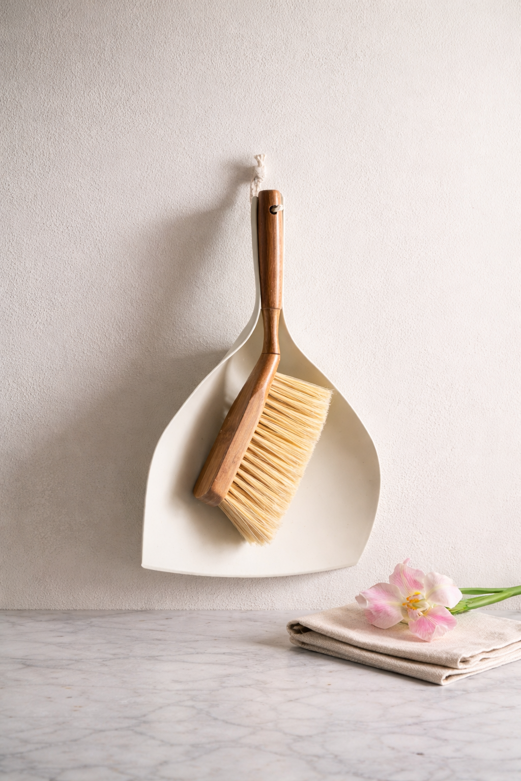 A cleaning brush with a wooden handle resting on a white dustpan, hanging on a wall, with a folded cloth and pink flowers on a marble surface below.