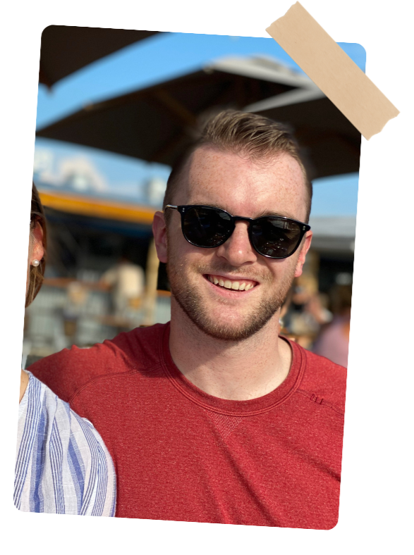 A young man with short light brown hair and a beard, wearing black sunglasses and a red t-shirt, smiling outdoors at a sunny location with a beach or boardwalk in the background.