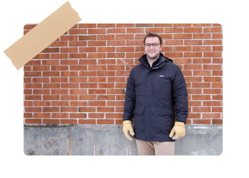 A man in a dark jacket, beige pants, and yellow gloves stands in front of a red brick wall, smiling.