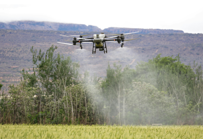 An agricultural drone flying over a green field with trees and mountains in the background.