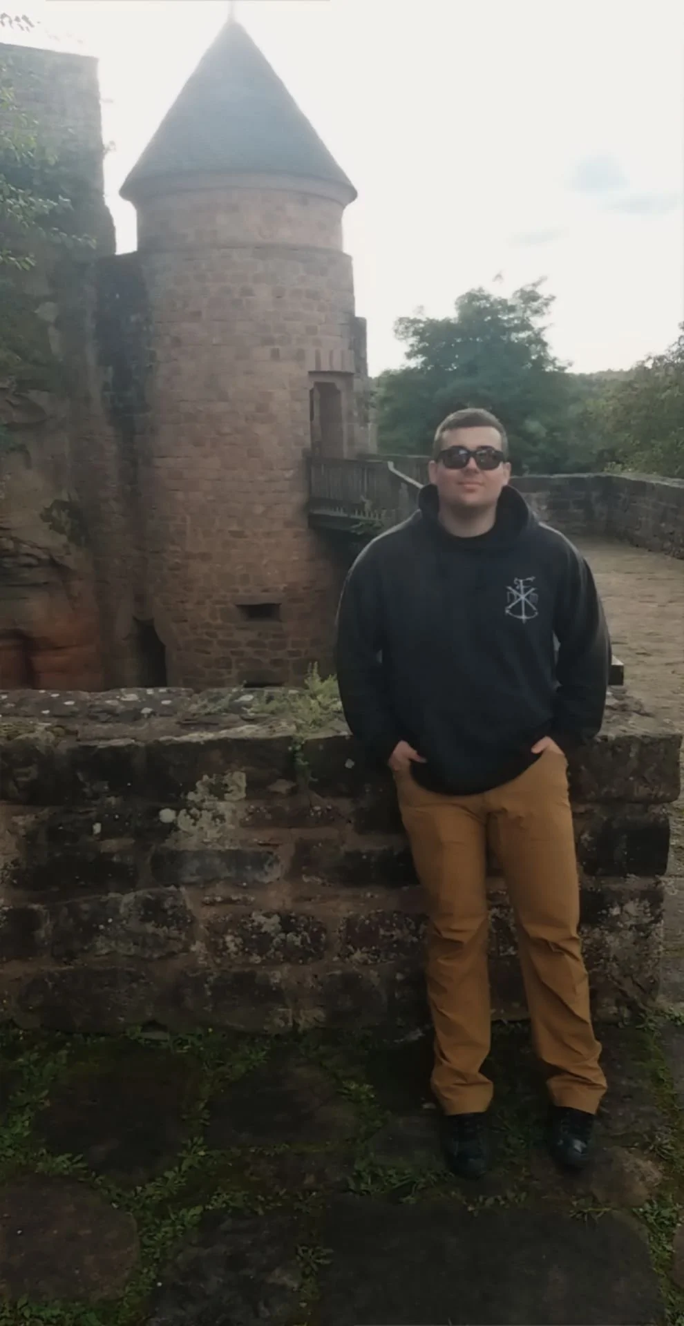 A young man wearing sunglasses, a black hoodie, and tan pants standing outdoors in front of a stone wall and a castle with a tower.