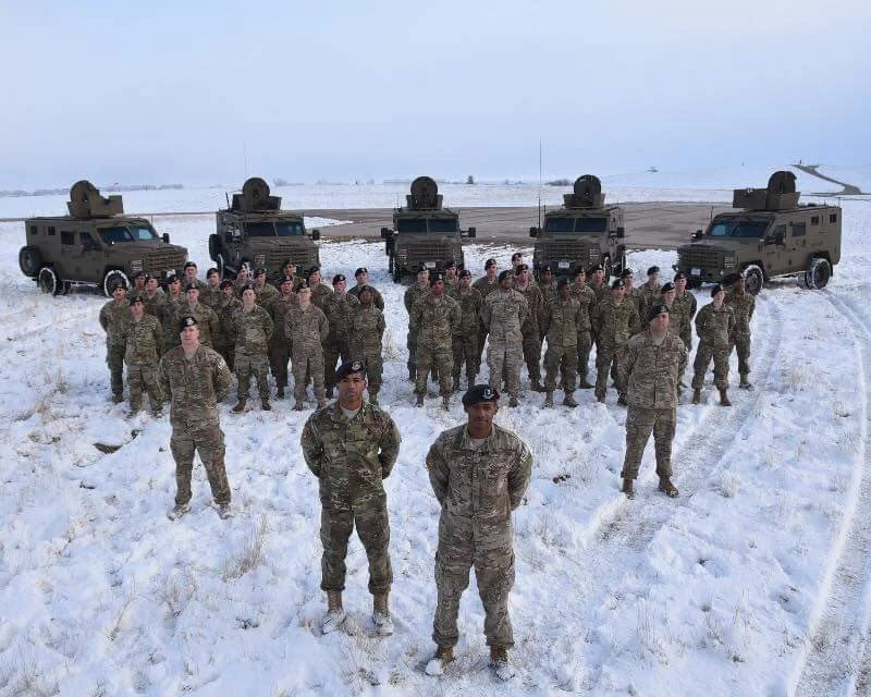 Group of soldiers in camouflage uniforms standing on snow with military vehicles in the background.