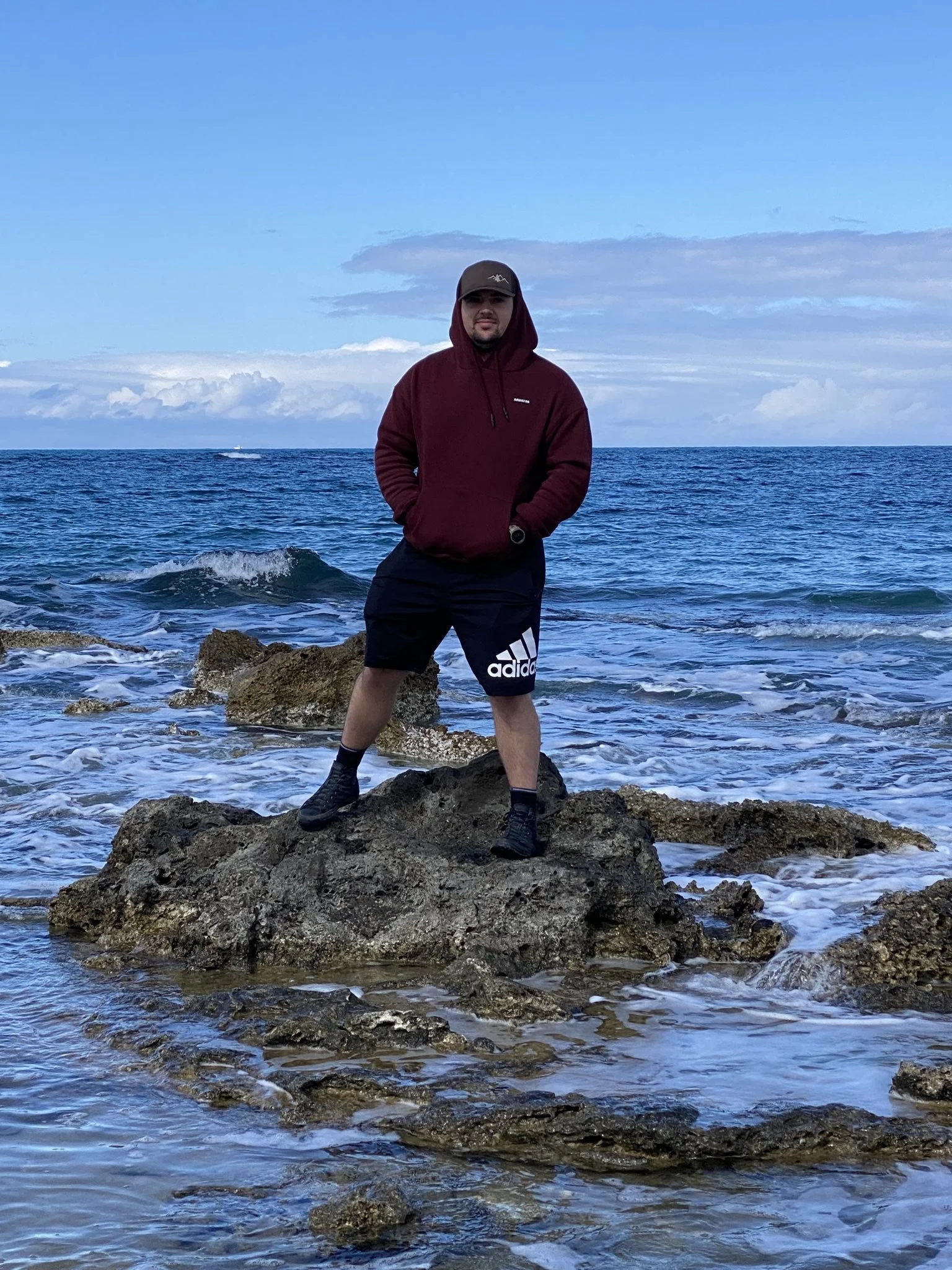 A man wearing a maroon hoodie, black Adidas shorts, and a gray cap stands on a large rock in the ocean with waves crashing around. The sky is partly cloudy with a blue backdrop.