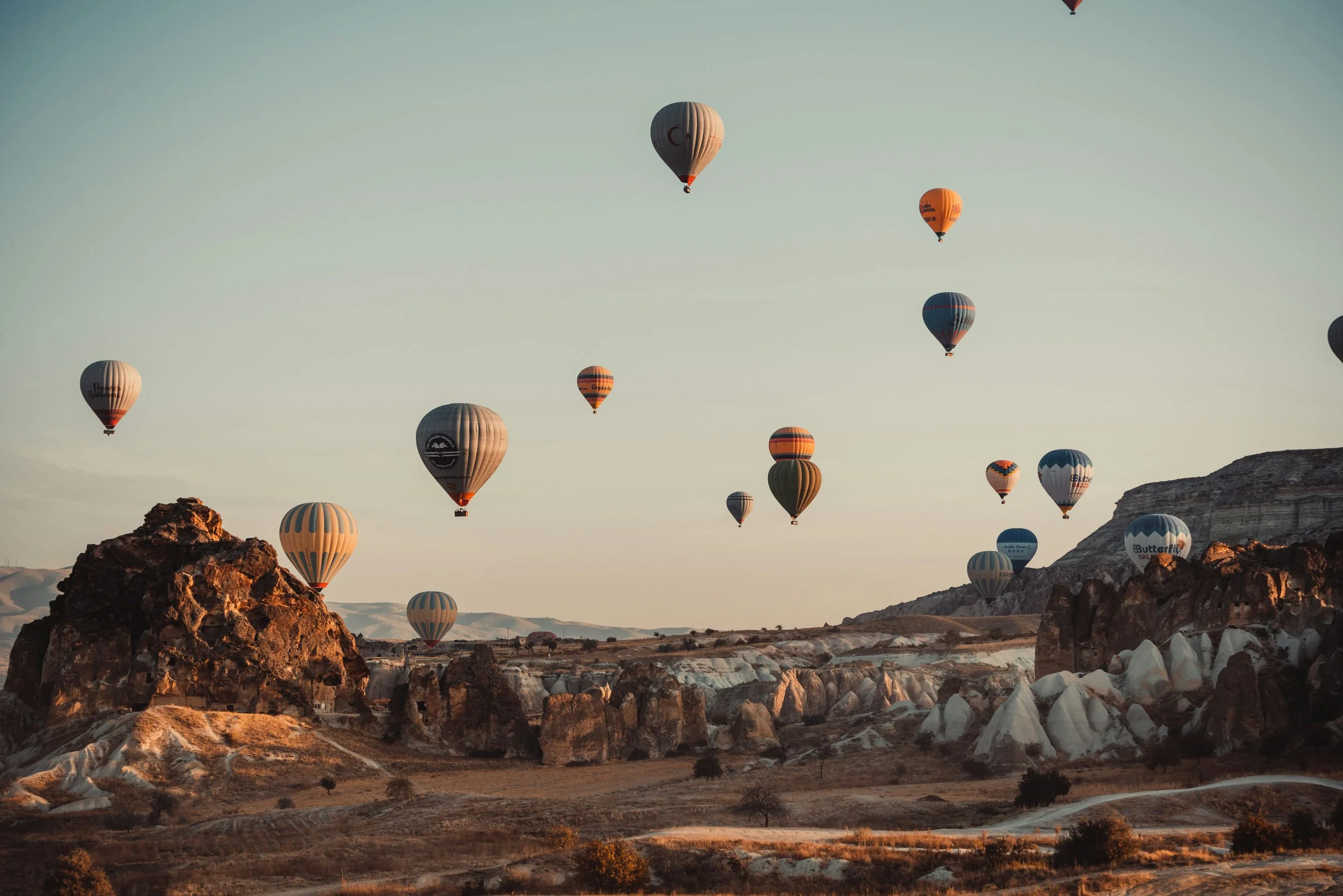 Multiple hot air balloons floating in the sky over a rocky landscape at sunrise or sunset, with some balloons near the ground and uneven terrain.