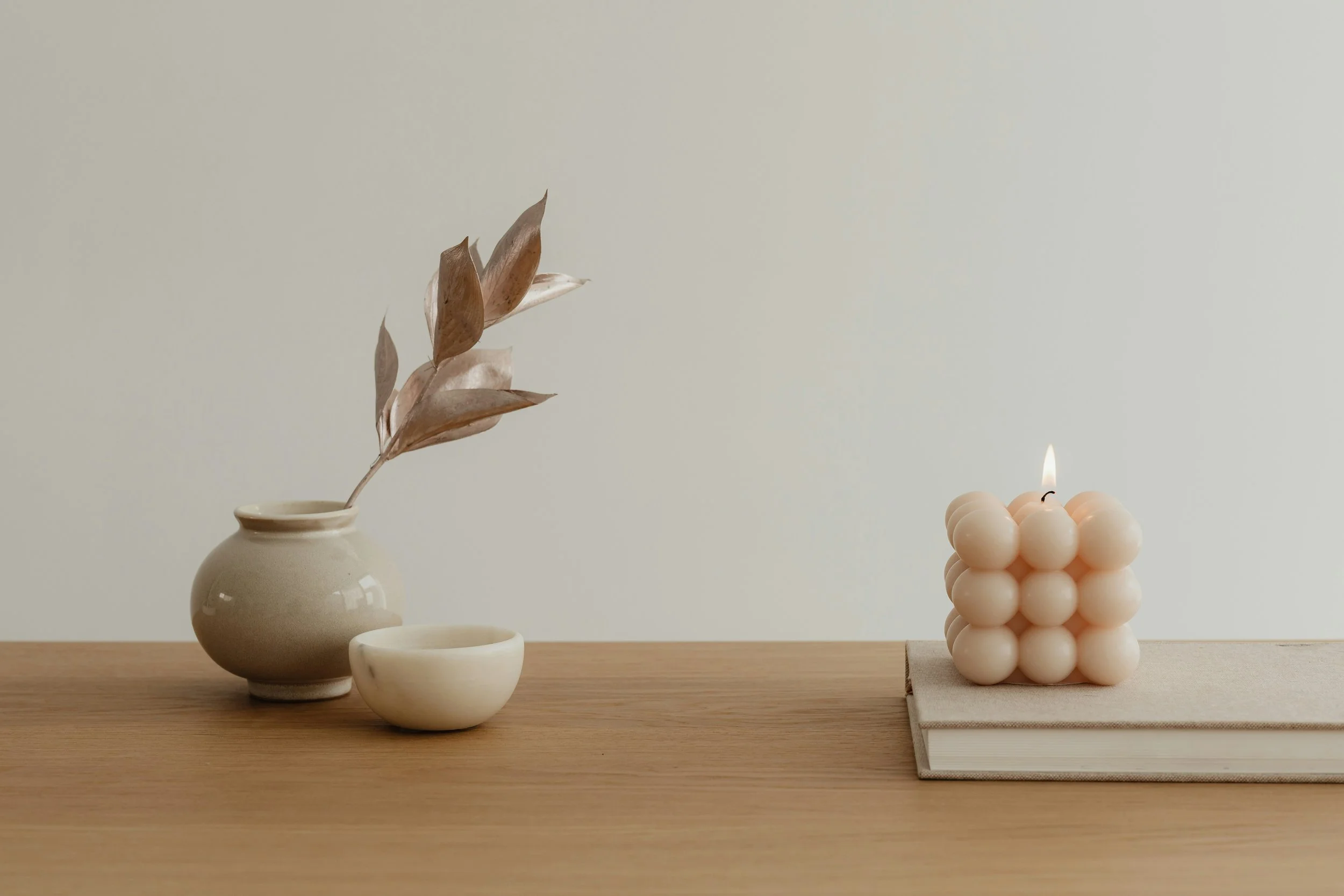 A minimalist still life featuring a white vase with dried leaves, three small white bowls, and a layered candle on a wooden surface against a plain, light wall background.