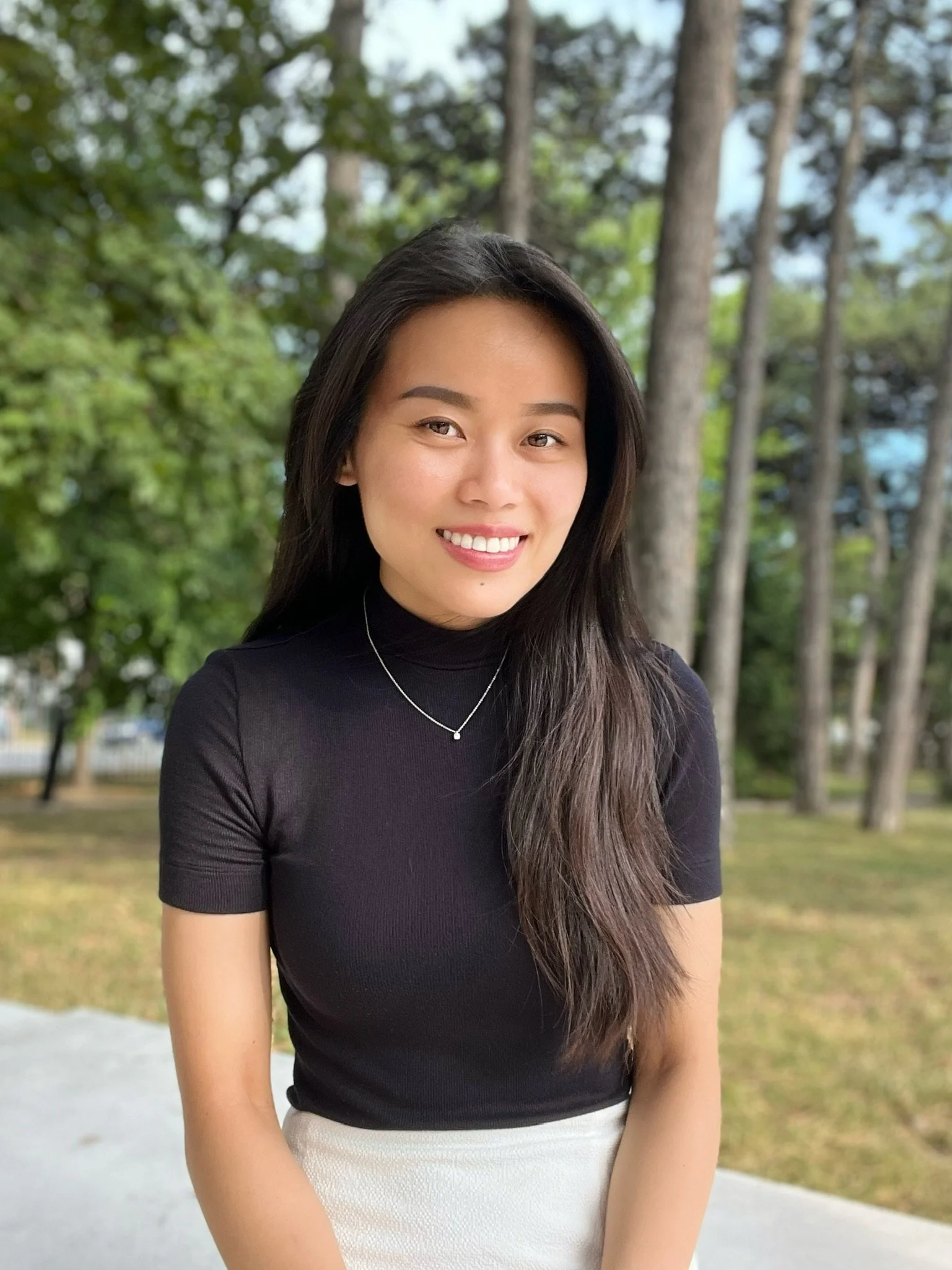 A young woman with long dark hair smiling outdoors, wearing a black top, with a background of trees and greenery.