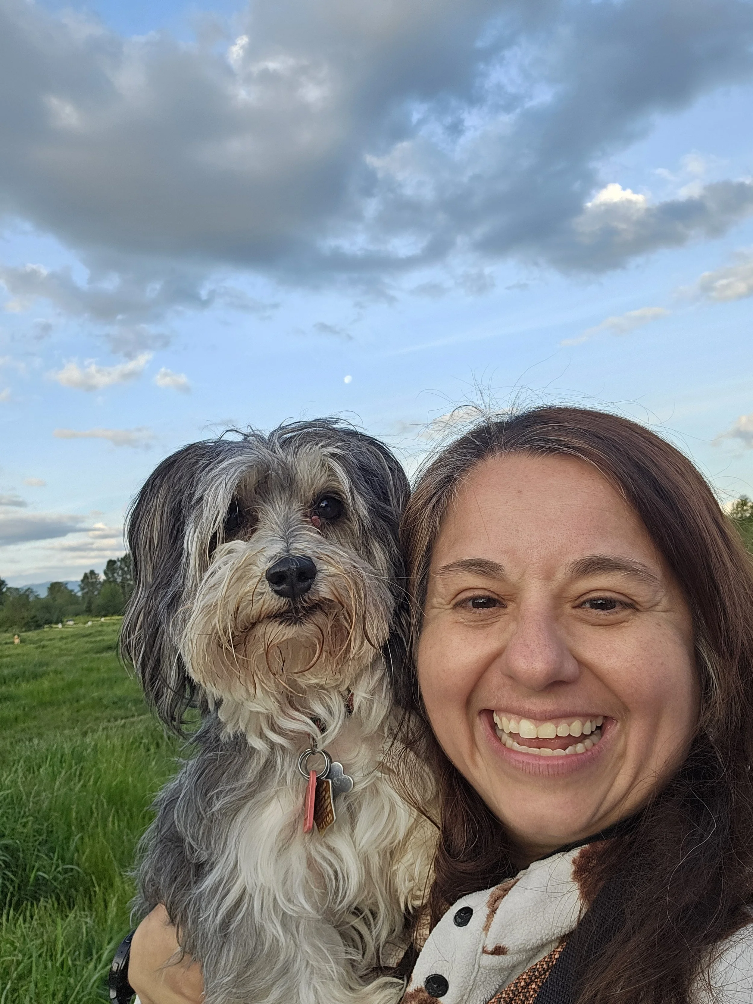 A woman smiling happily while holding a small, fluffy dog outdoors in a green field under a partly cloudy sky with the moon visible.