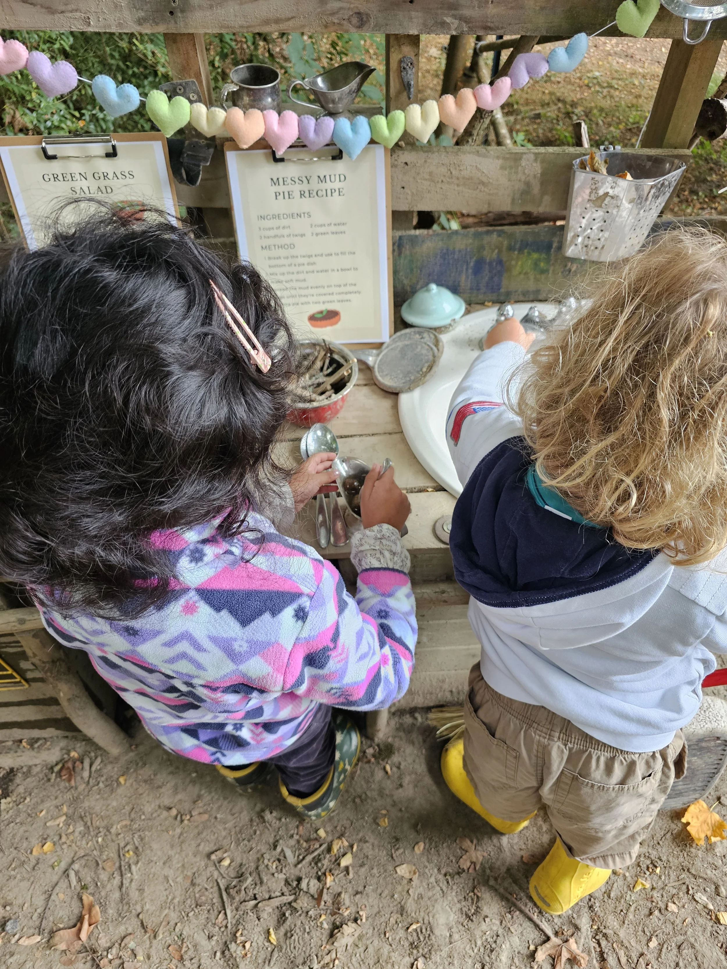 Two children are at a rustic outdoor play kitchen. One girl with dark curly hair, wearing a colorful patterned jacket, is holding kitchen utensils. The other child, with blond curly hair, in a gray hoodie, beige shorts, and yellow rain boots, is interacting with the play setup. Above, there are signs with recipes and a decorative string of pastel-colored heart-shaped ornaments.