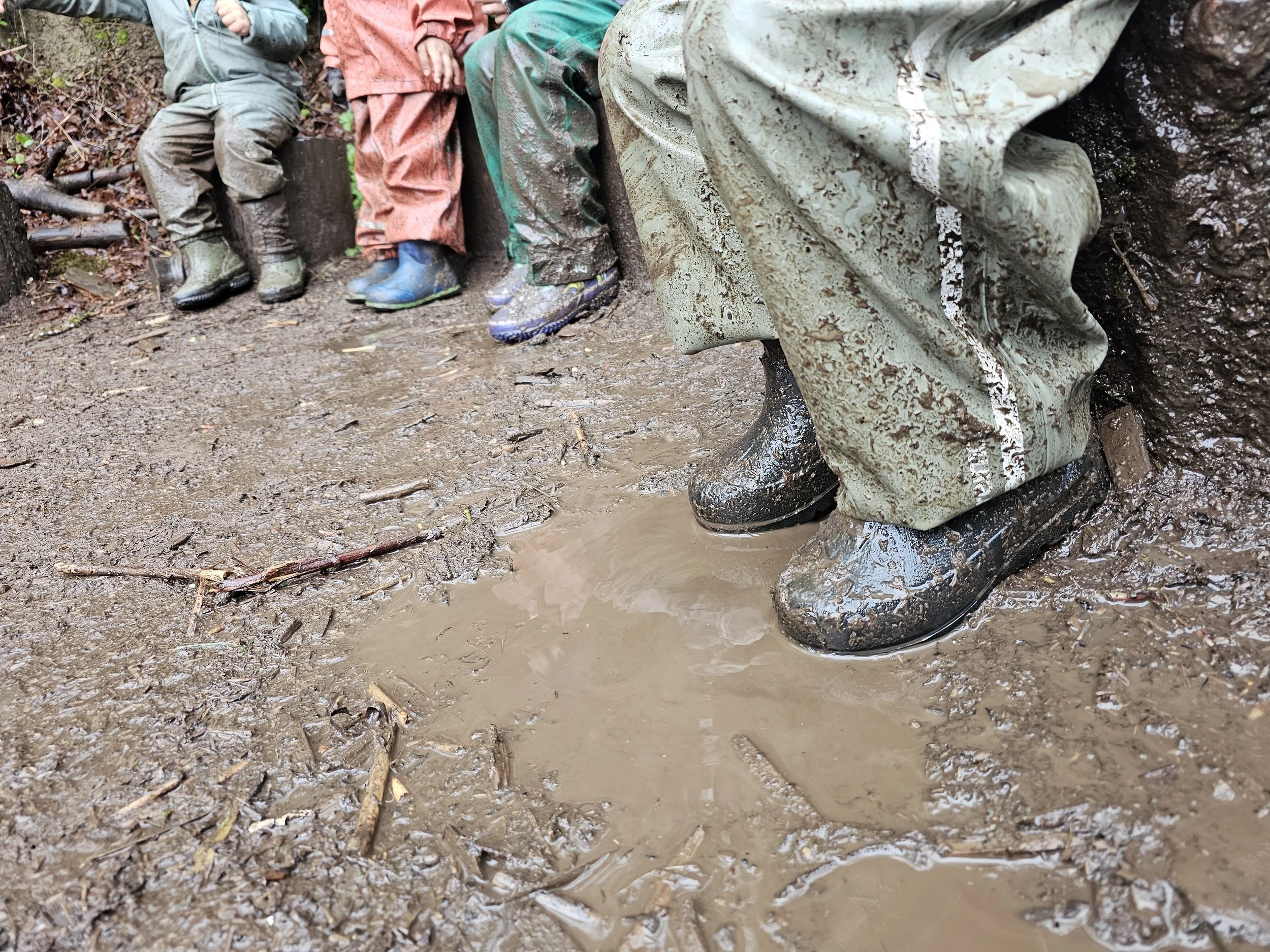 Children sitting on a muddy ground wearing boots, with muddy boots and wet rain gear visible.