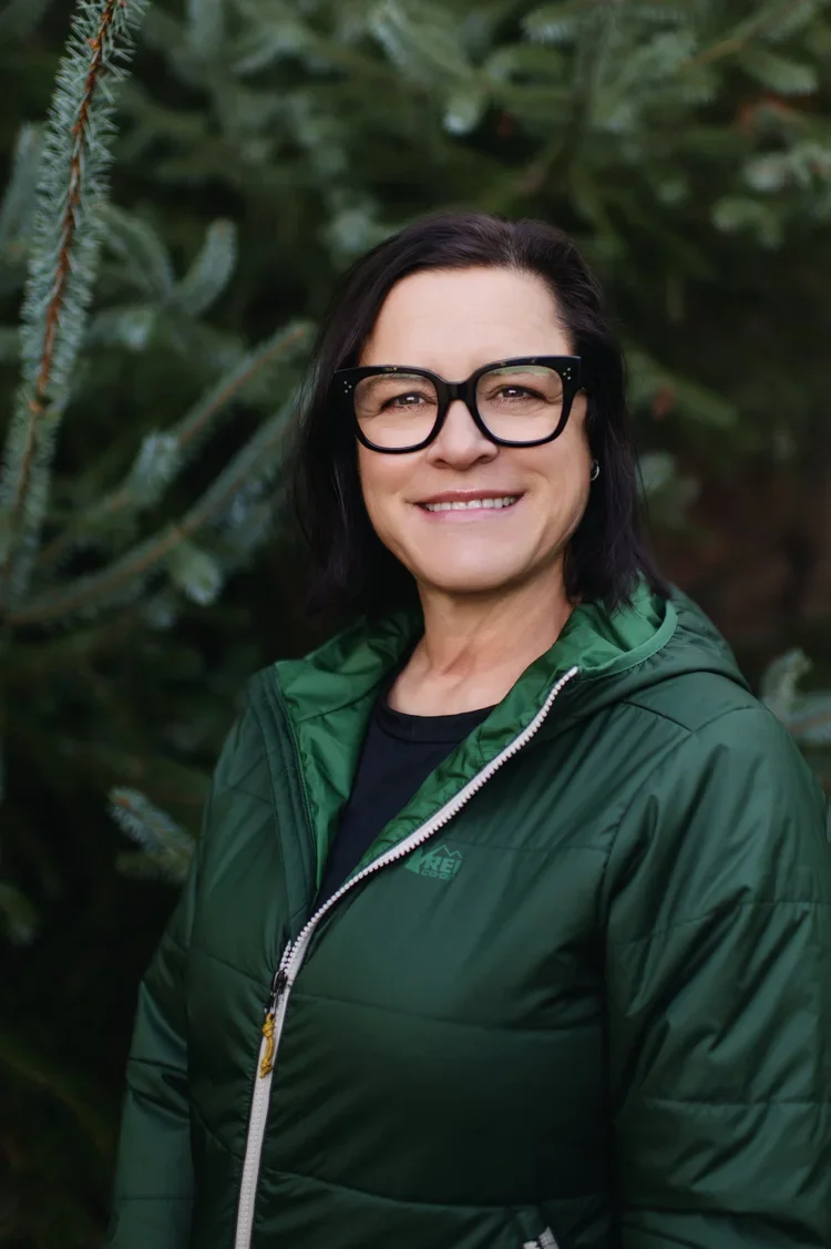 A woman with black hair and glasses smiling outdoors in front of green pine trees, wearing a green jacket.