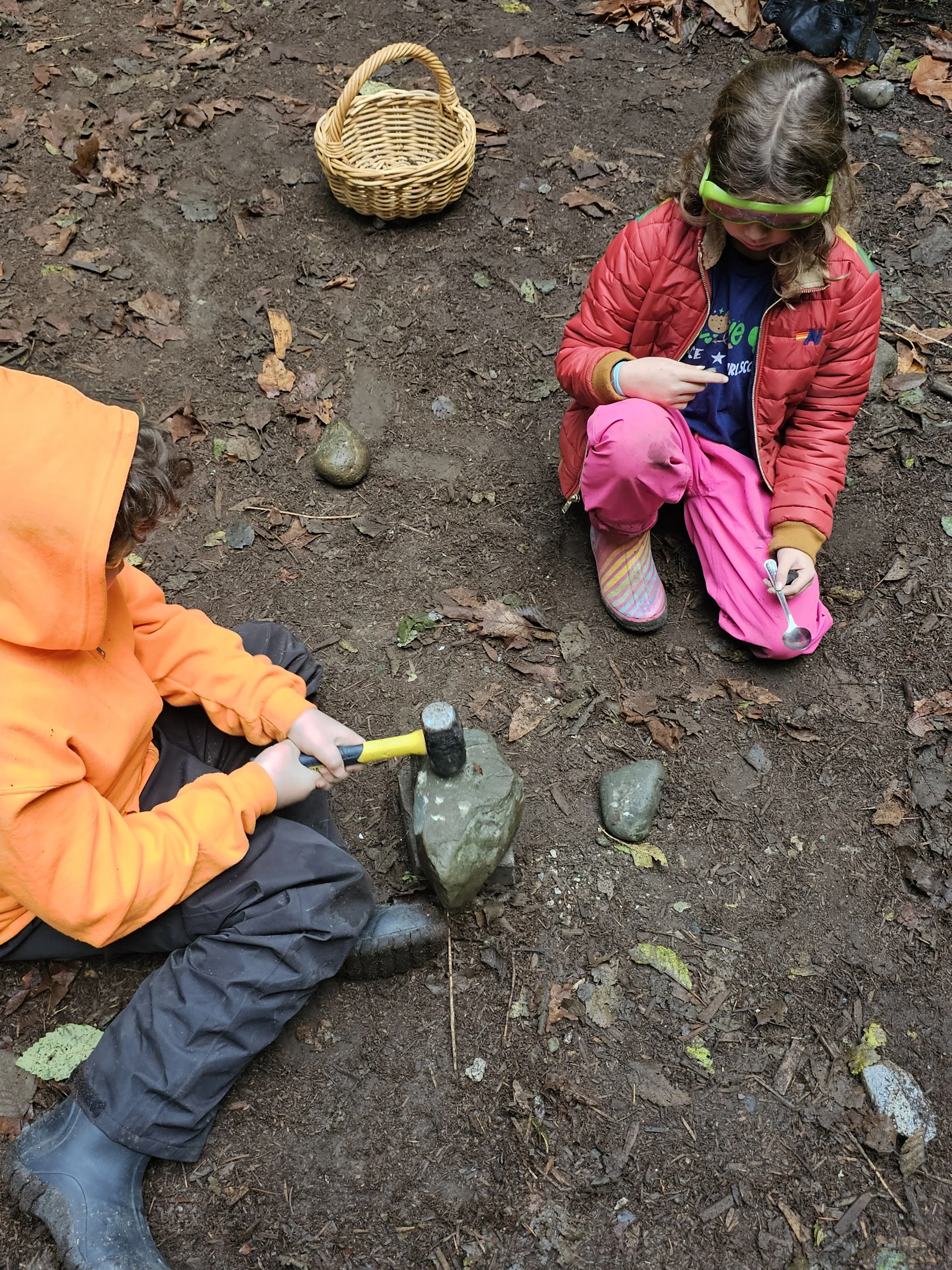 Two children outdoors on dirt ground with scattered leaves. One child in an orange jacket is hitting a rock with a hammer, and the other child in a red jacket and pink pants is kneeling nearby holding a small spoon.