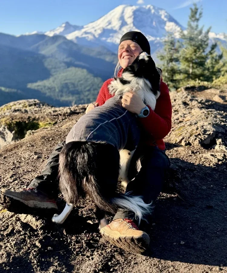 A person in outdoor gear sitting on rocky ground, hugging two black-and-white dogs, with a snow-capped mountain and green trees in the background.