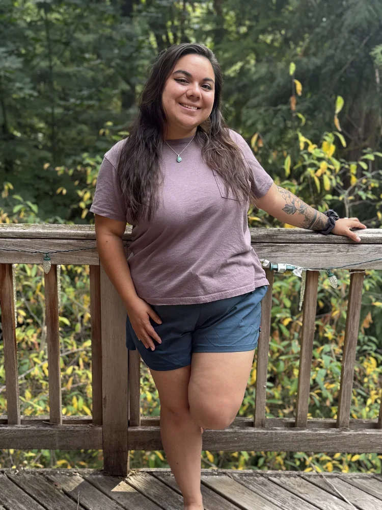 A woman with long dark hair, smiling, standing on a wooden deck outdoors, wearing a mauve T-shirt and dark blue shorts, with a tattoo on her right arm, surrounded by green trees.