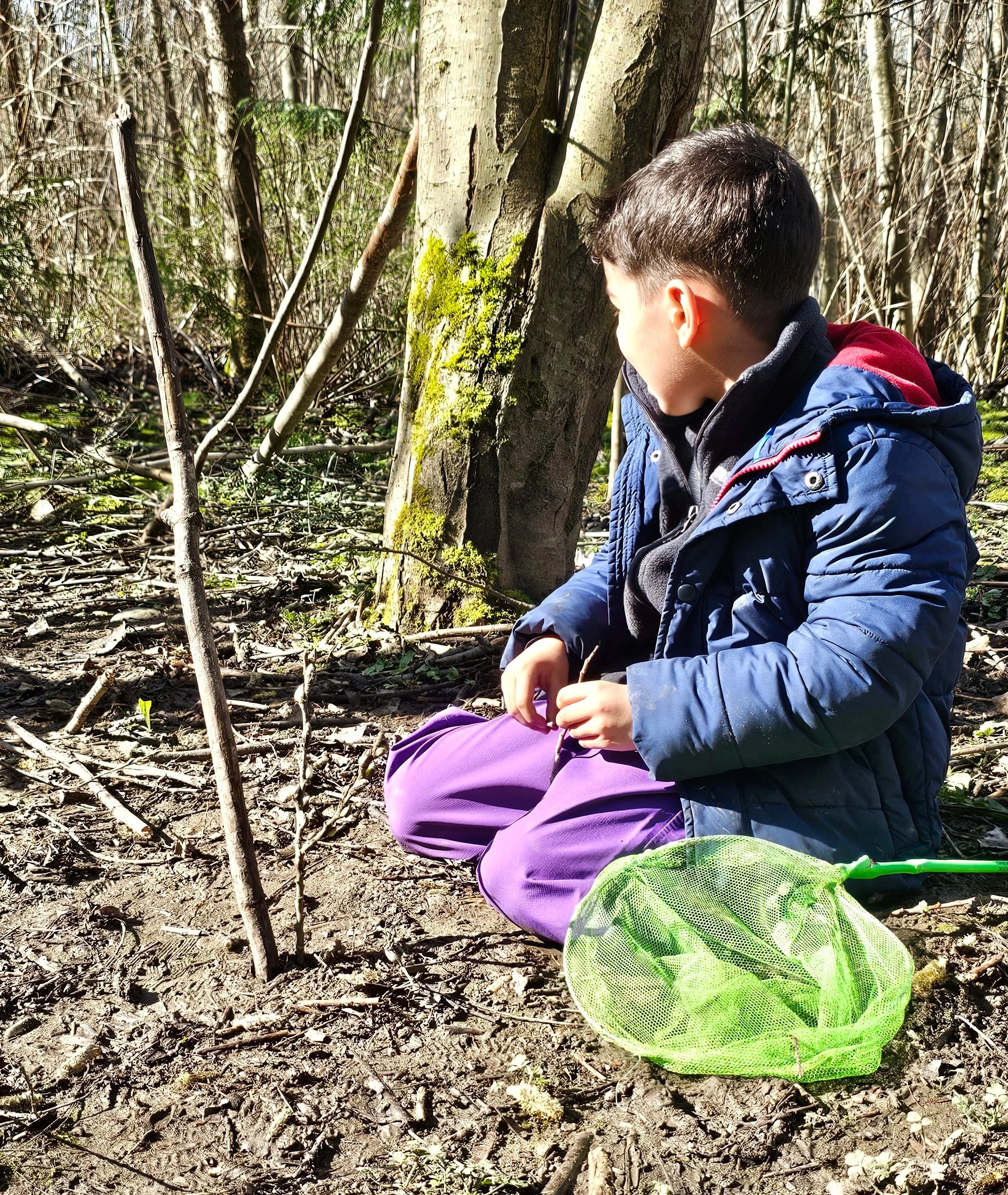 A boy sitting cross-legged in a forest, looking at trees and plants, with a green net in front of him.