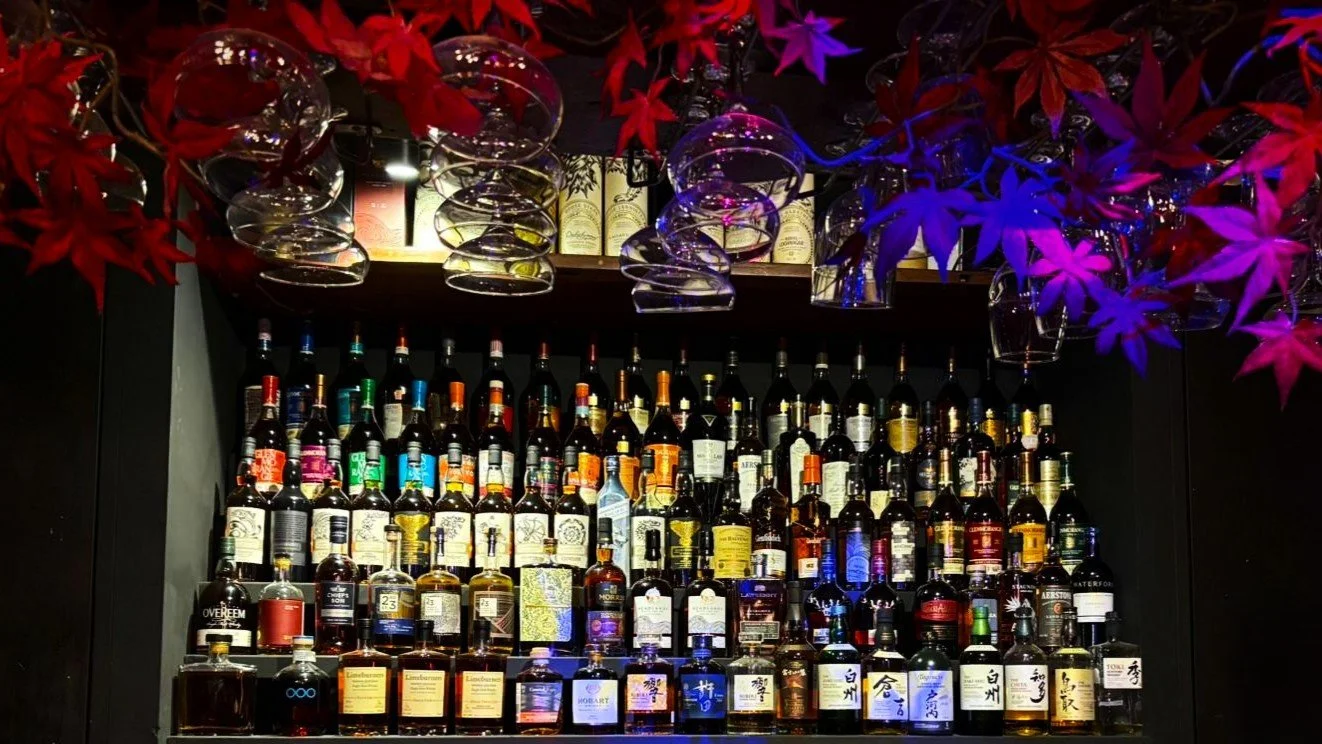 A well-stocked bar with a variety of liquor bottles, purple and red decorative leaves, and upside-down wine glasses hanging from above.