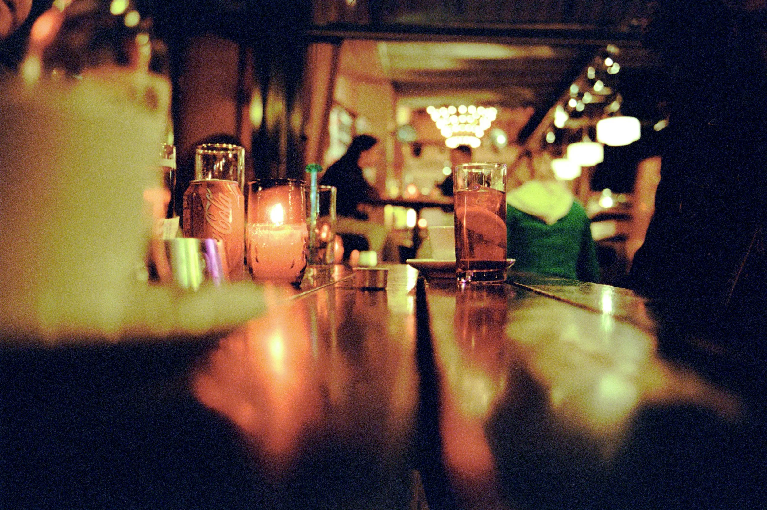 Dimly lit bar or restaurant with drinks on the table, including a can of Coca-Cola and glasses of water, and blurred background of patrons and decorative lighting.