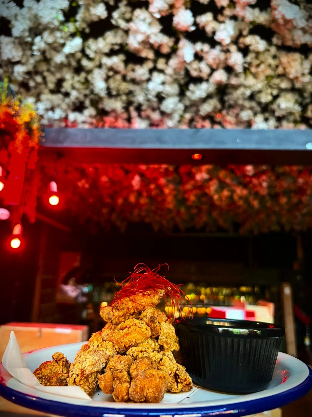 Plate of fried chicken pieces garnished with red chili threads, served with a black dipping sauce container, in a restaurant with colorful flower or paper decoration on the ceiling.
