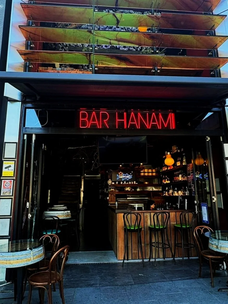 The entrance of a bar named Bar Hanam, with a bright red neon sign above the door. There are outdoor tables and chairs in front of the bar, and inside, shelves with bottles and a bar counter are visible.