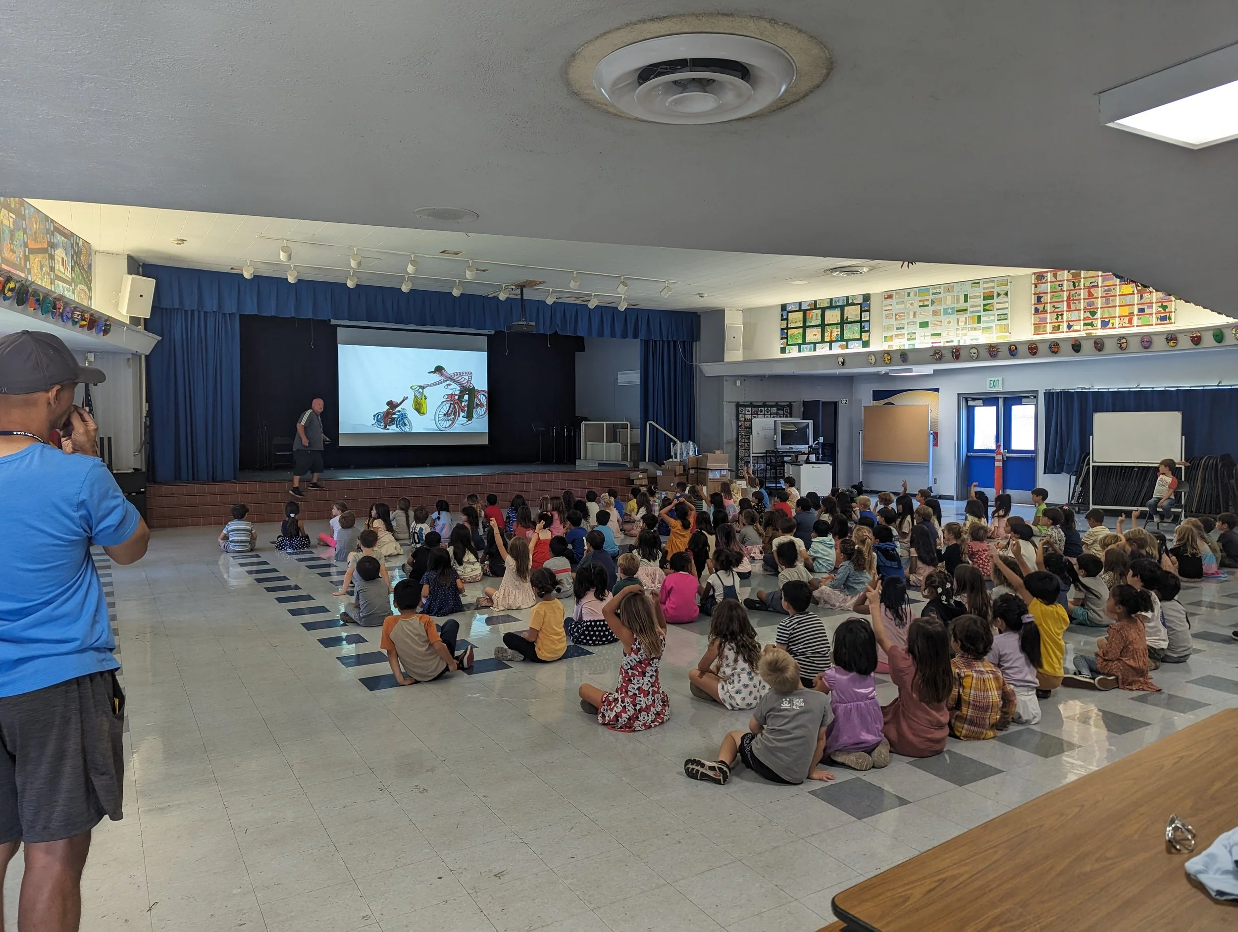 Many children sitting on the floor in an auditorium watching a video on the projector, using the AV system to sync Audio and Visual for the kids.